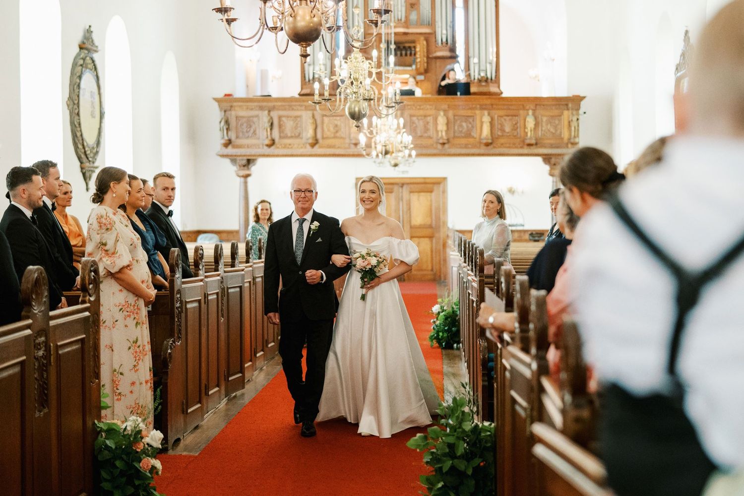 Bride walks down church aisle with father on red carpet, guests standing in wooden pews during wedding ceremony.