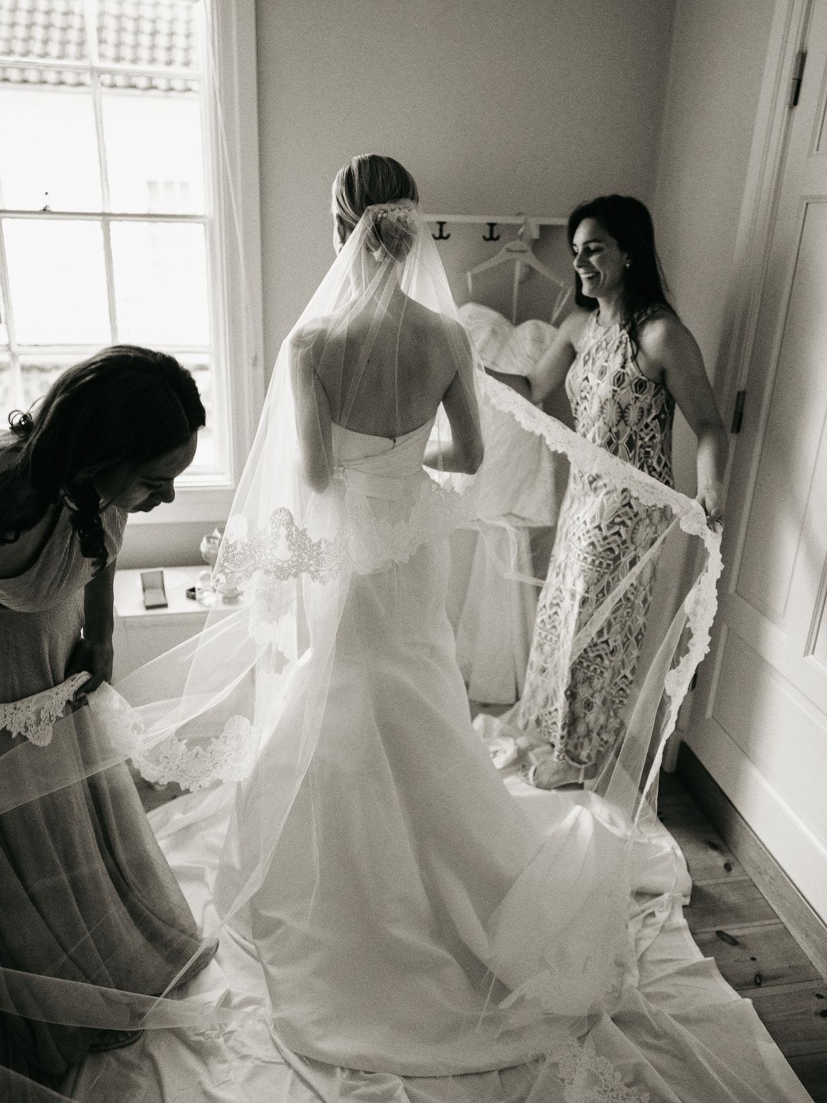 Black and white photo of a bride in a wedding gown and veil being assisted by two bridesmaids before the ceremony.