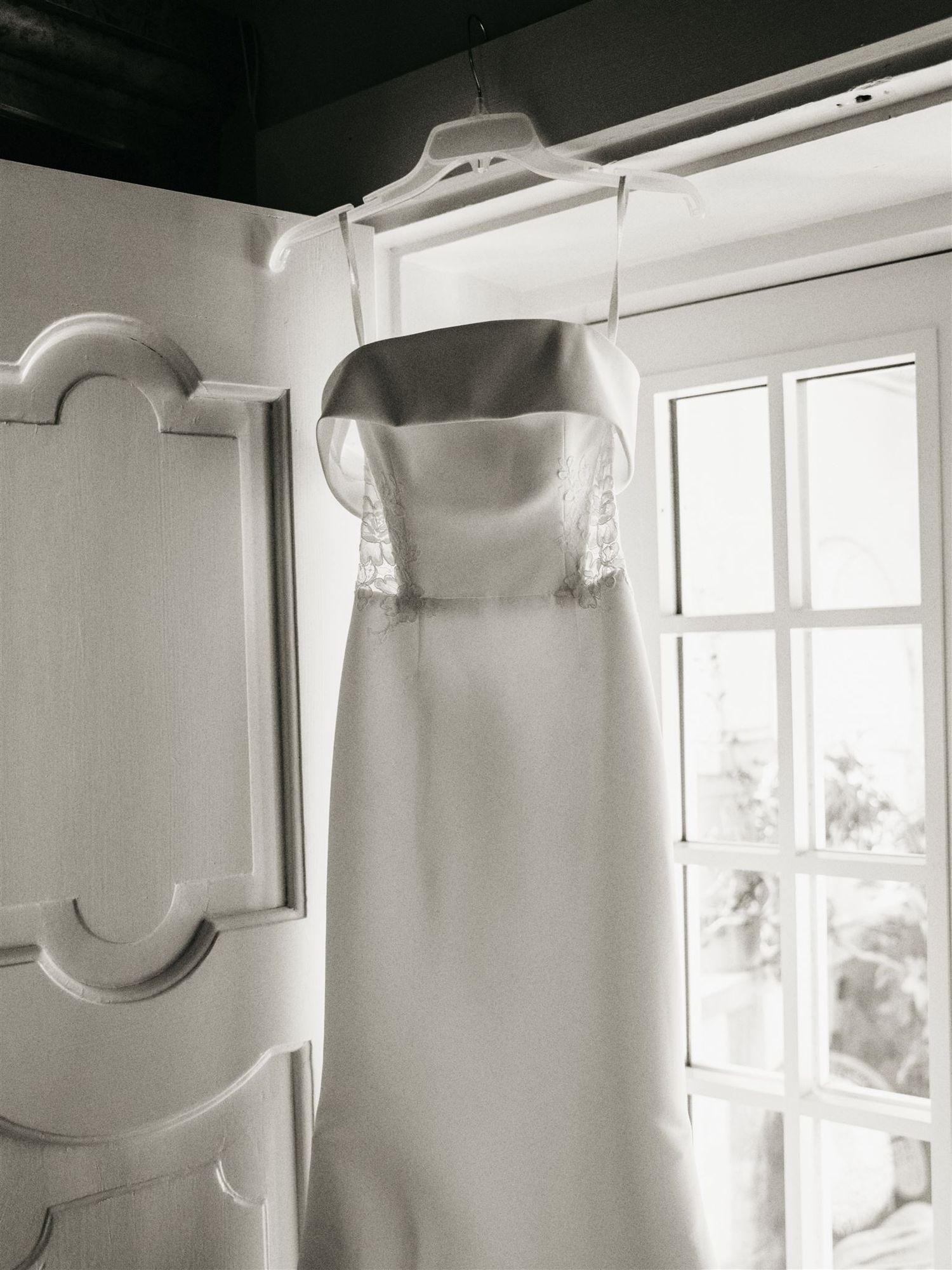 Elegant strapless white wedding gown hanging in a window in black and white photography.