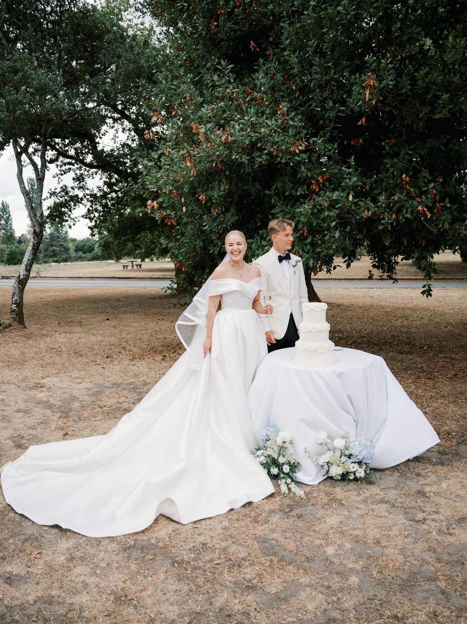 Bride and groom posing beside elegant three-tier white wedding cake under a tree at outdoor chateau wedding.