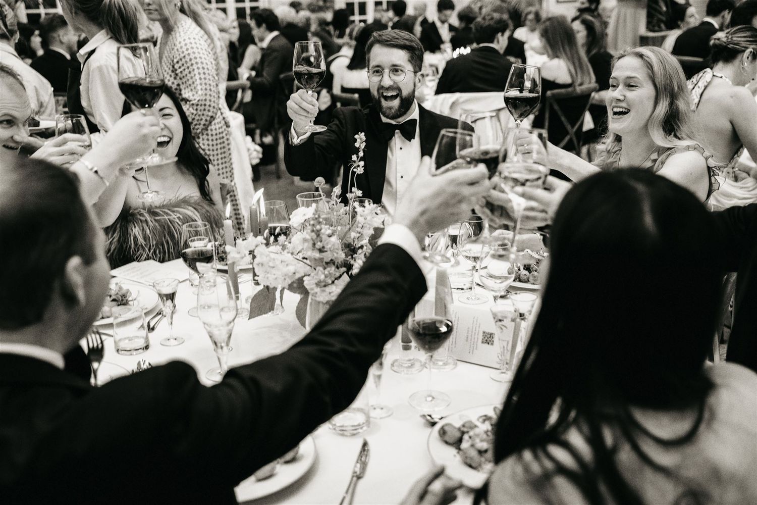 Black and white photo of wedding guests toasting with wine glasses at reception.
