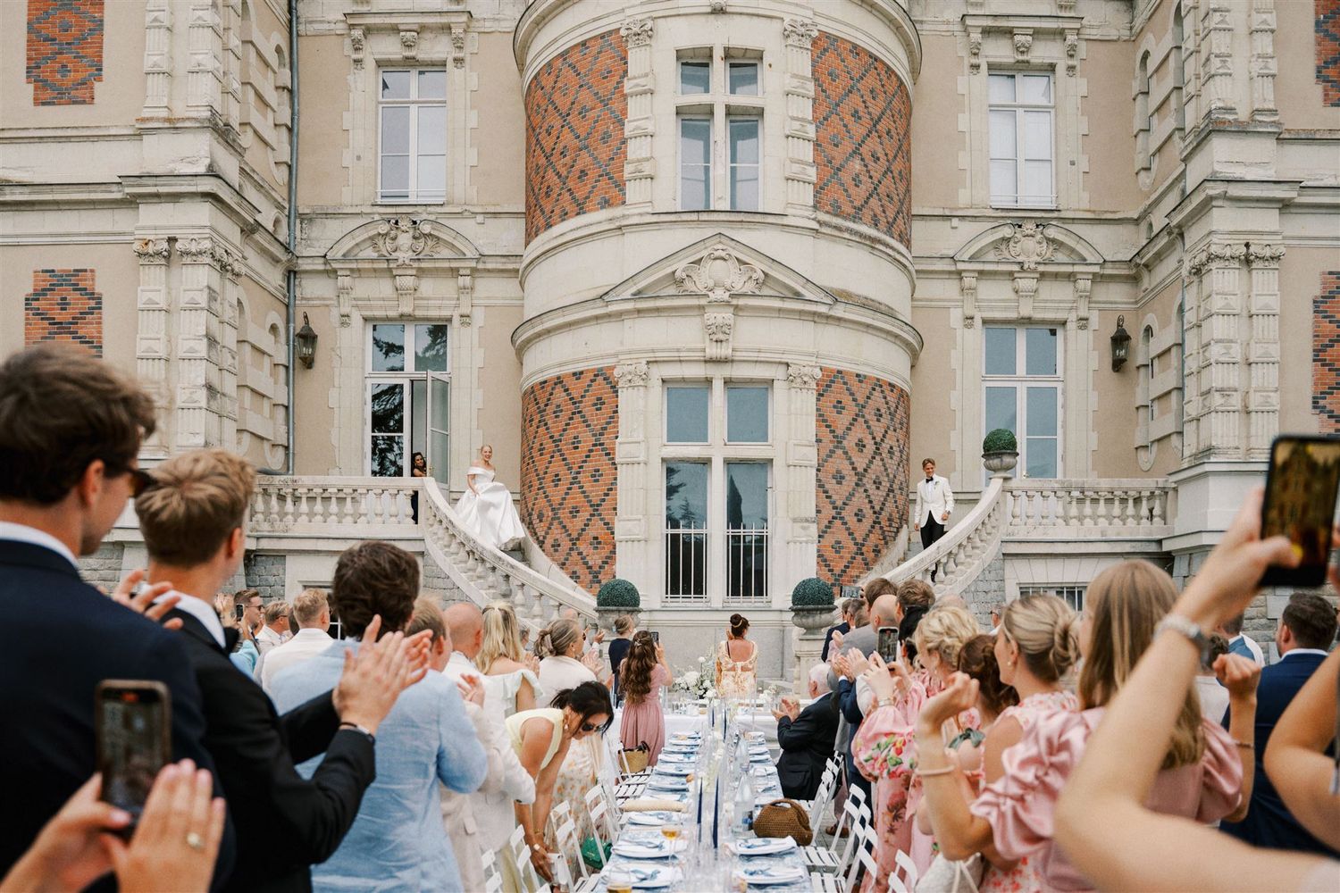 Wedding guests applaud couple at grand outdoor reception in front of ornate chateau facade.