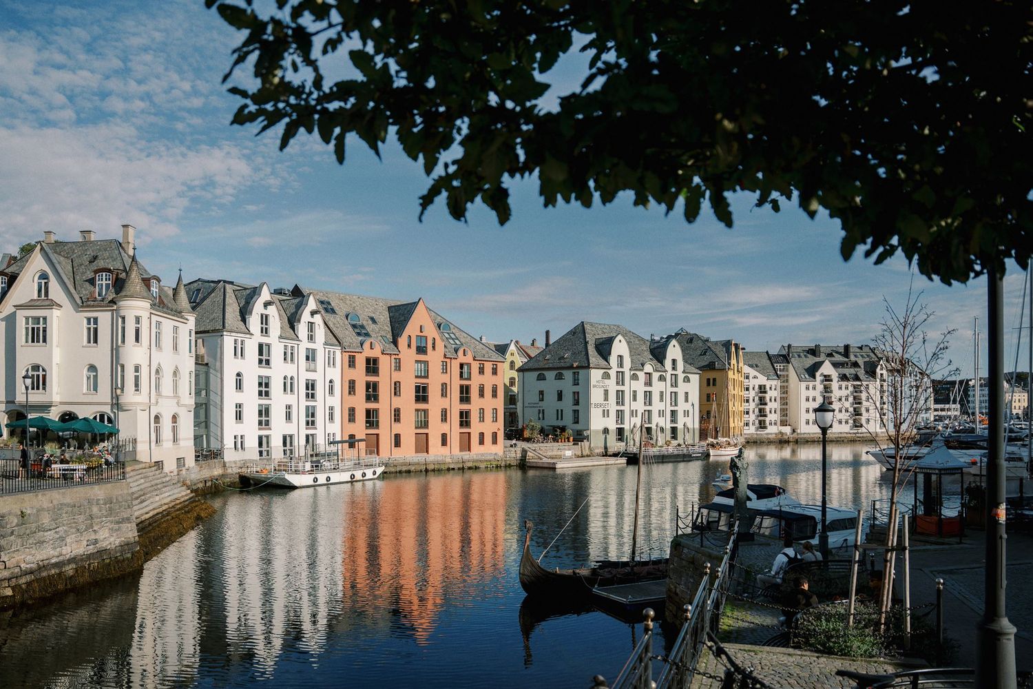 Colorful historic buildings reflecting in calm canal waters in Ålesund, Norway, under a partly cloudy sky.