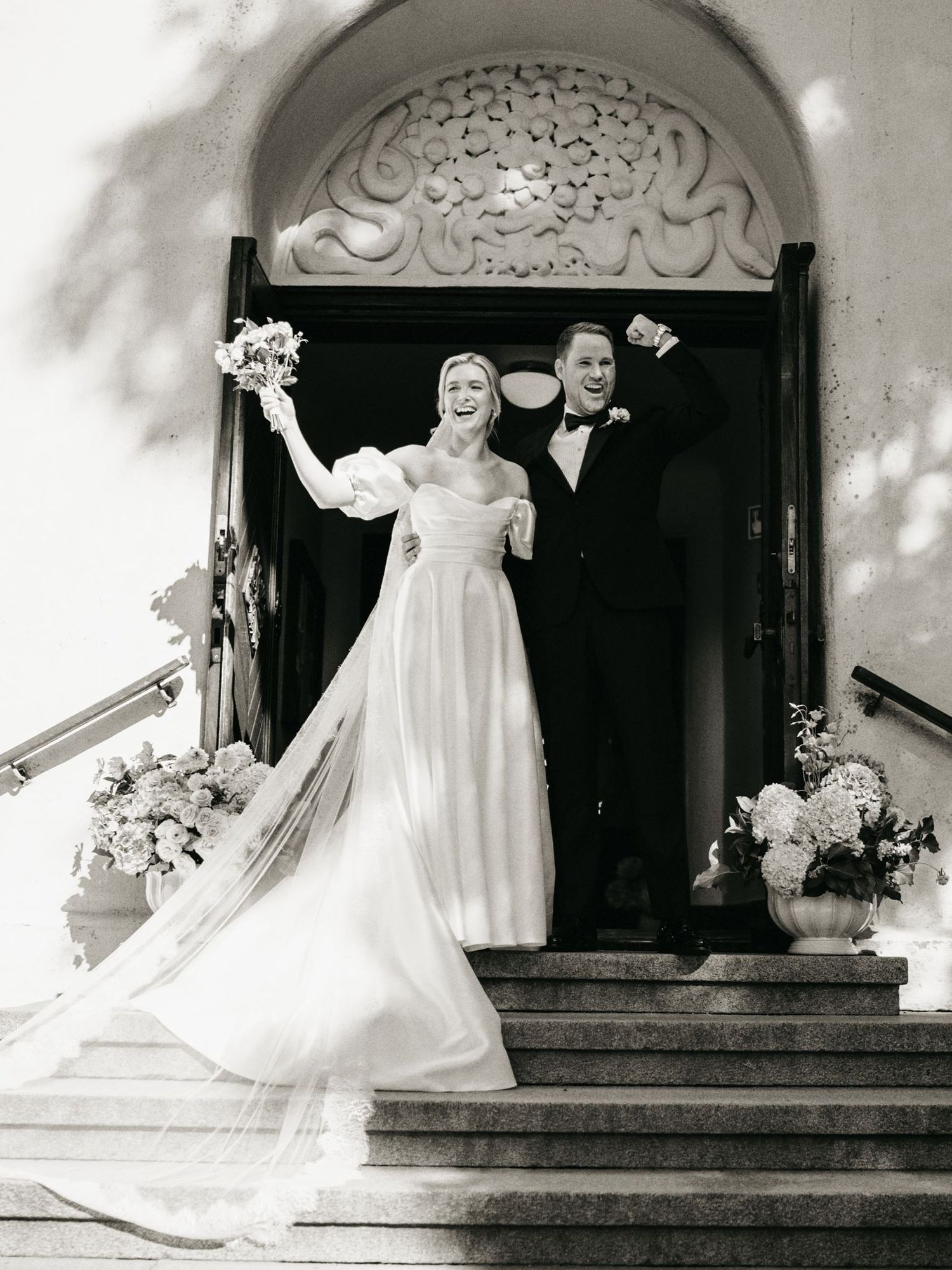 Black and white photo of joyful newlyweds celebrating on church steps, bride in flowing gown holding bouquet aloft.
