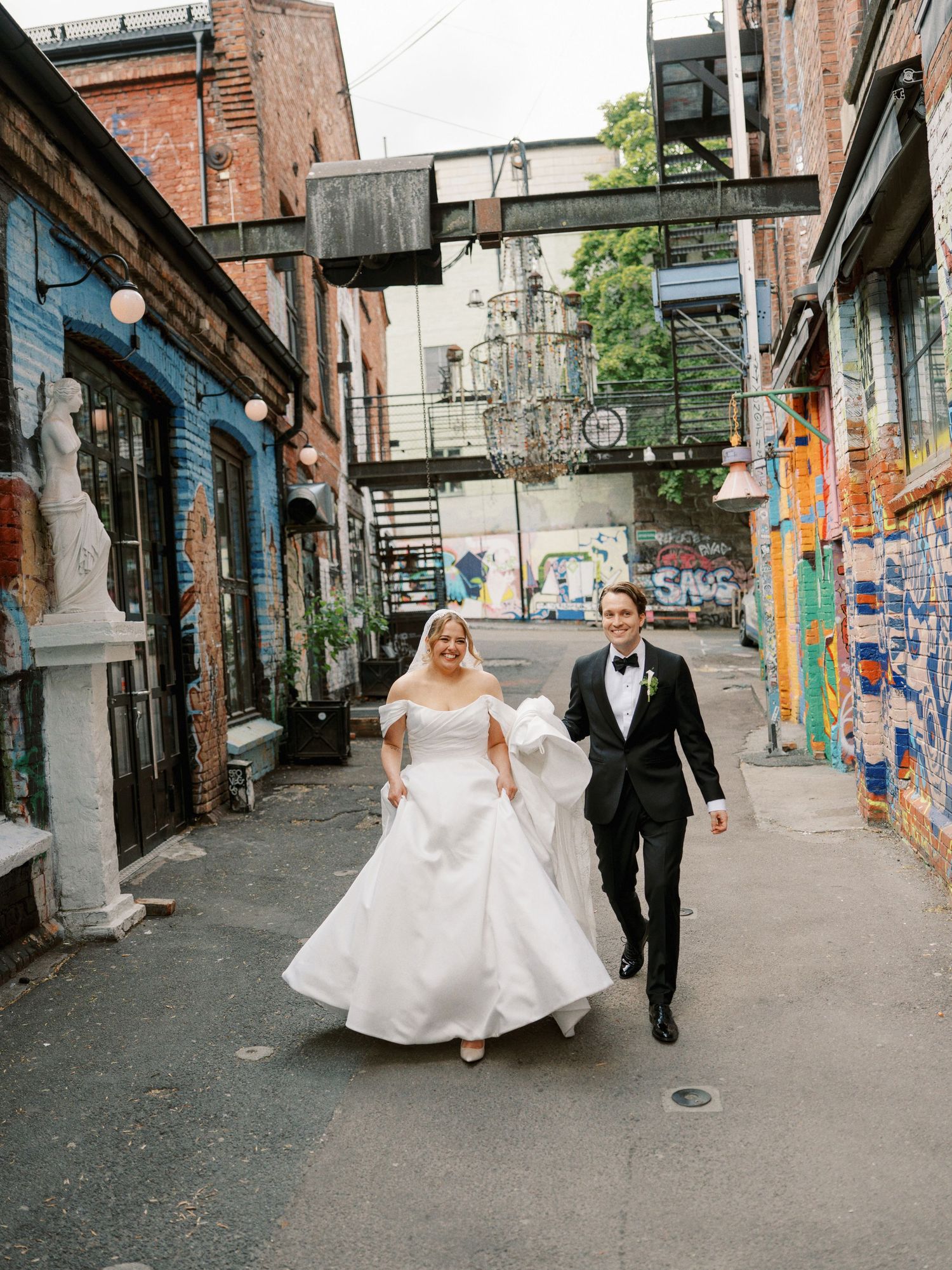 Bride and groom walk hand-in-hand through a colorful graffiti-lined alley on their wedding day.