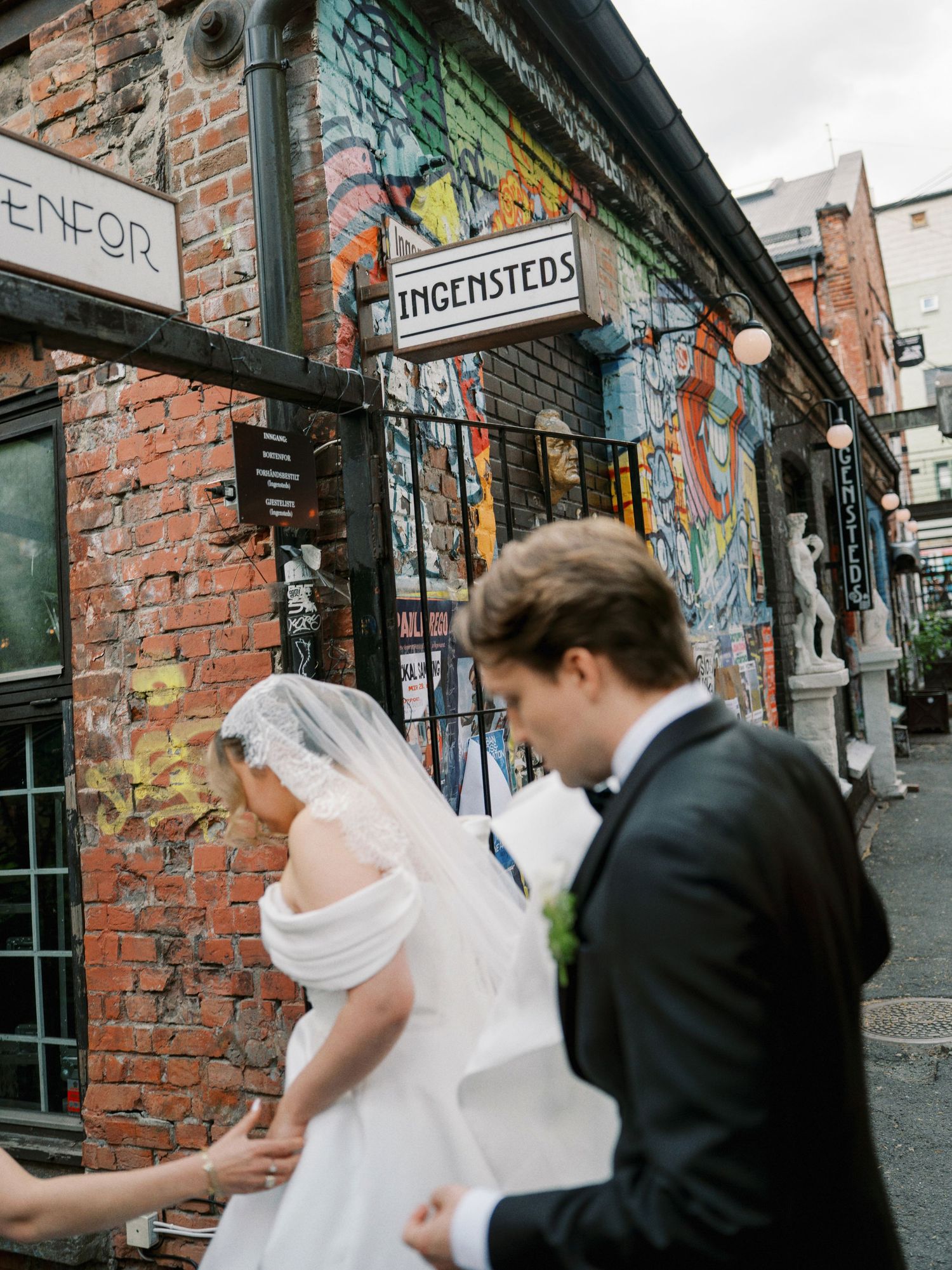 A bride and groom walk past a colorful graffiti-covered brick building with a sign reading Ingensteds.