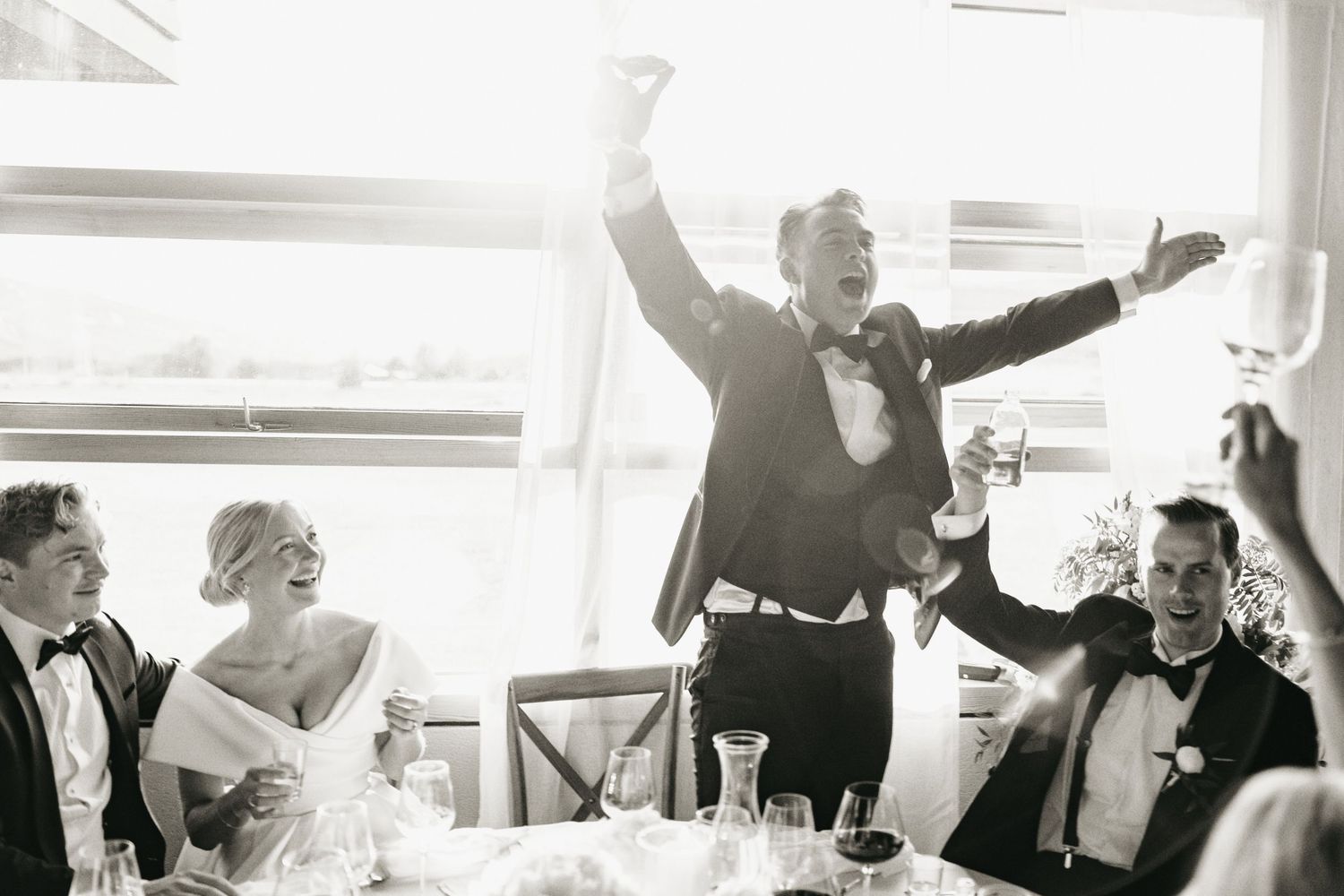 Groom celebrates joyfully with arms raised at wedding reception while bride and guests laugh at dinner table.