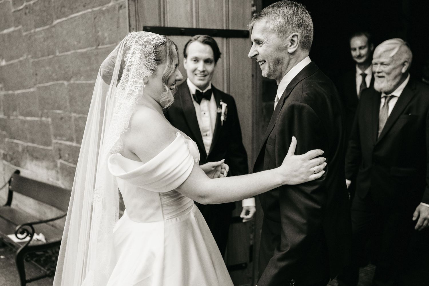 Black and white wedding photo of bride greeting guest while groom and groomsmen watch in elegant venue.