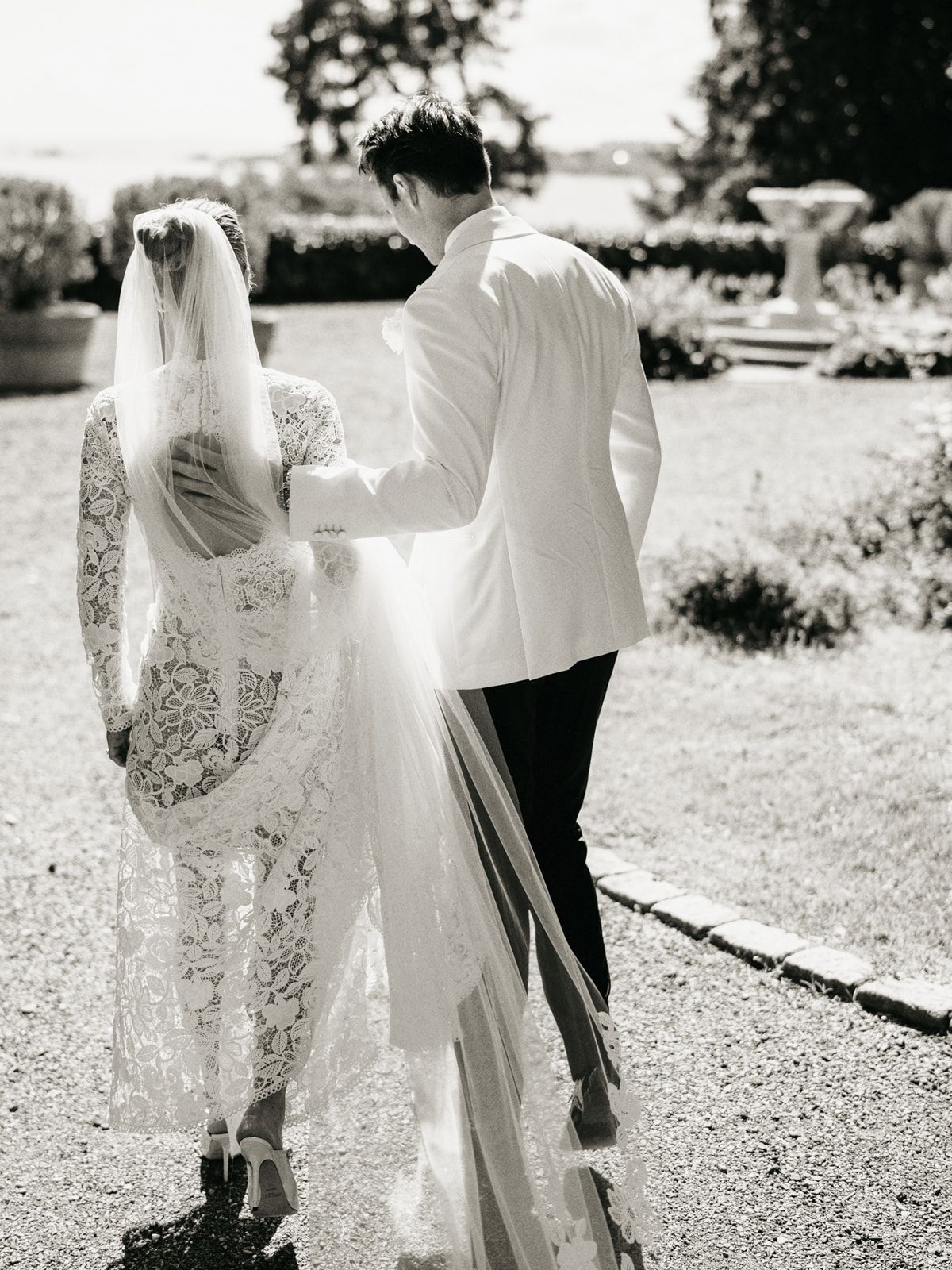 Black and white photo of bride in lace gown and veil walking with groom in white jacket through garden.