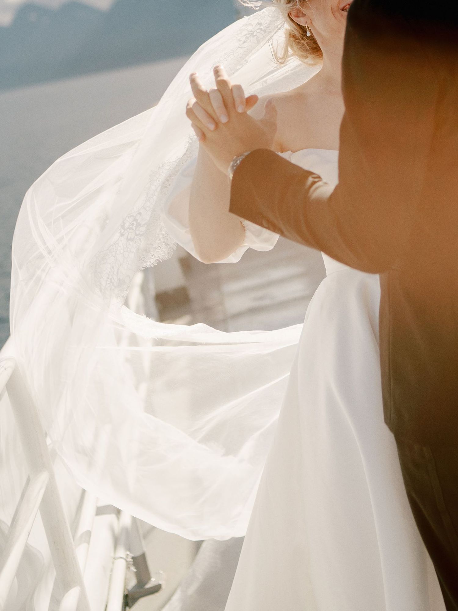Bride in flowing white wedding gown and veil being adjusted outdoors in soft sunlight near water.