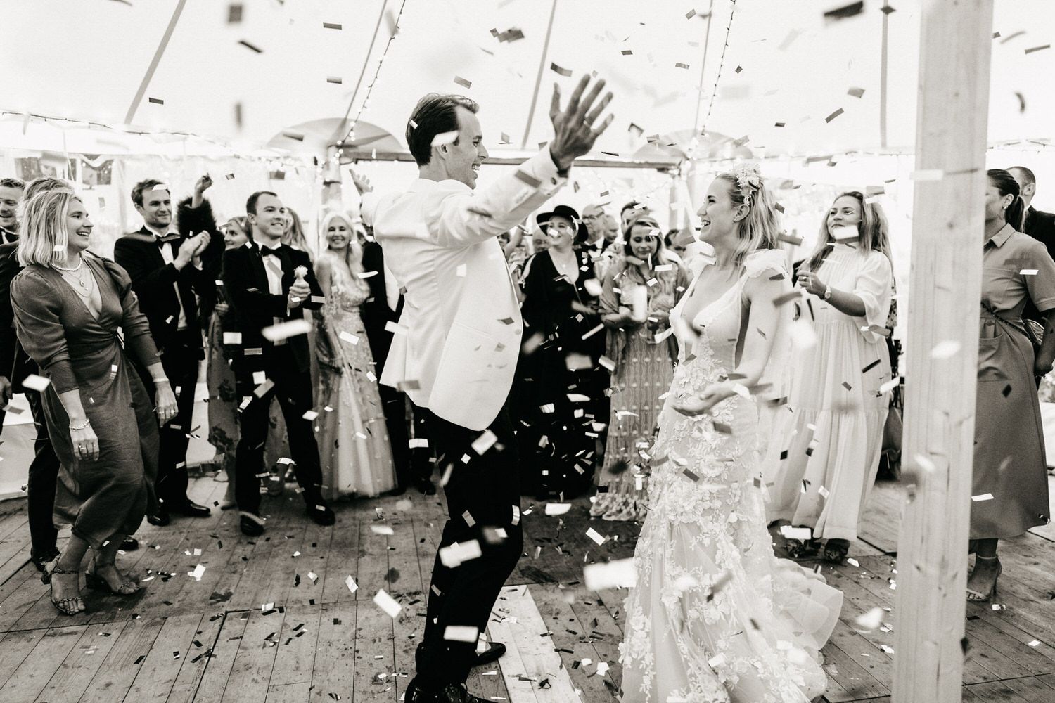 Black and white photo of bride and groom celebrating with confetti under a tent surrounded by cheering guests.