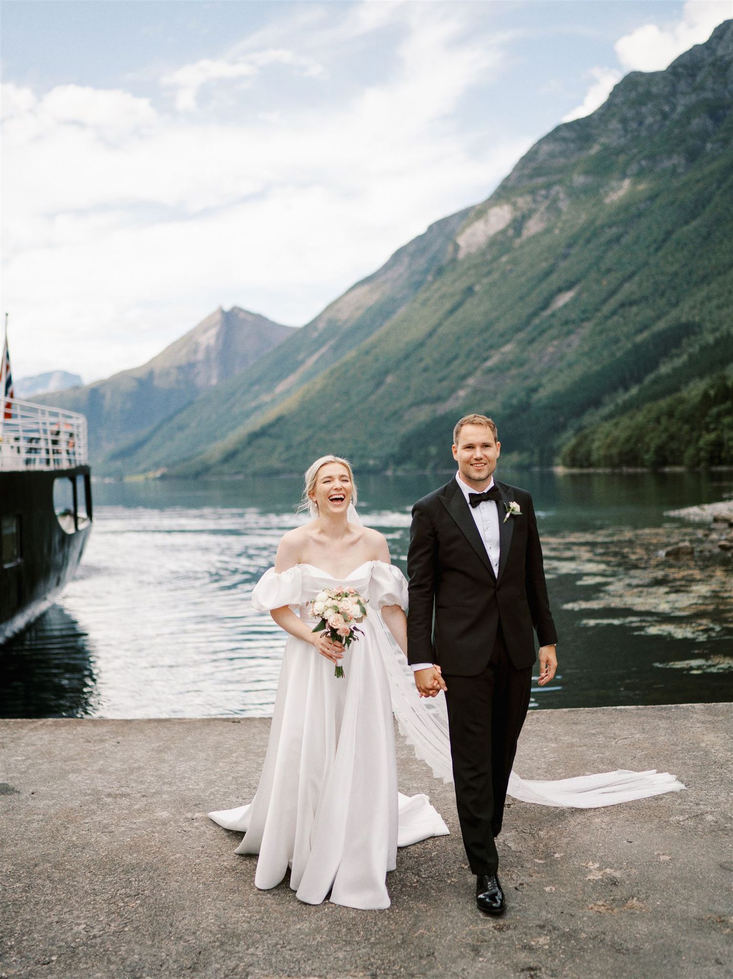 Laughing newlyweds stroll along fjord dock with stunning mountain and lake scenery in Norway.