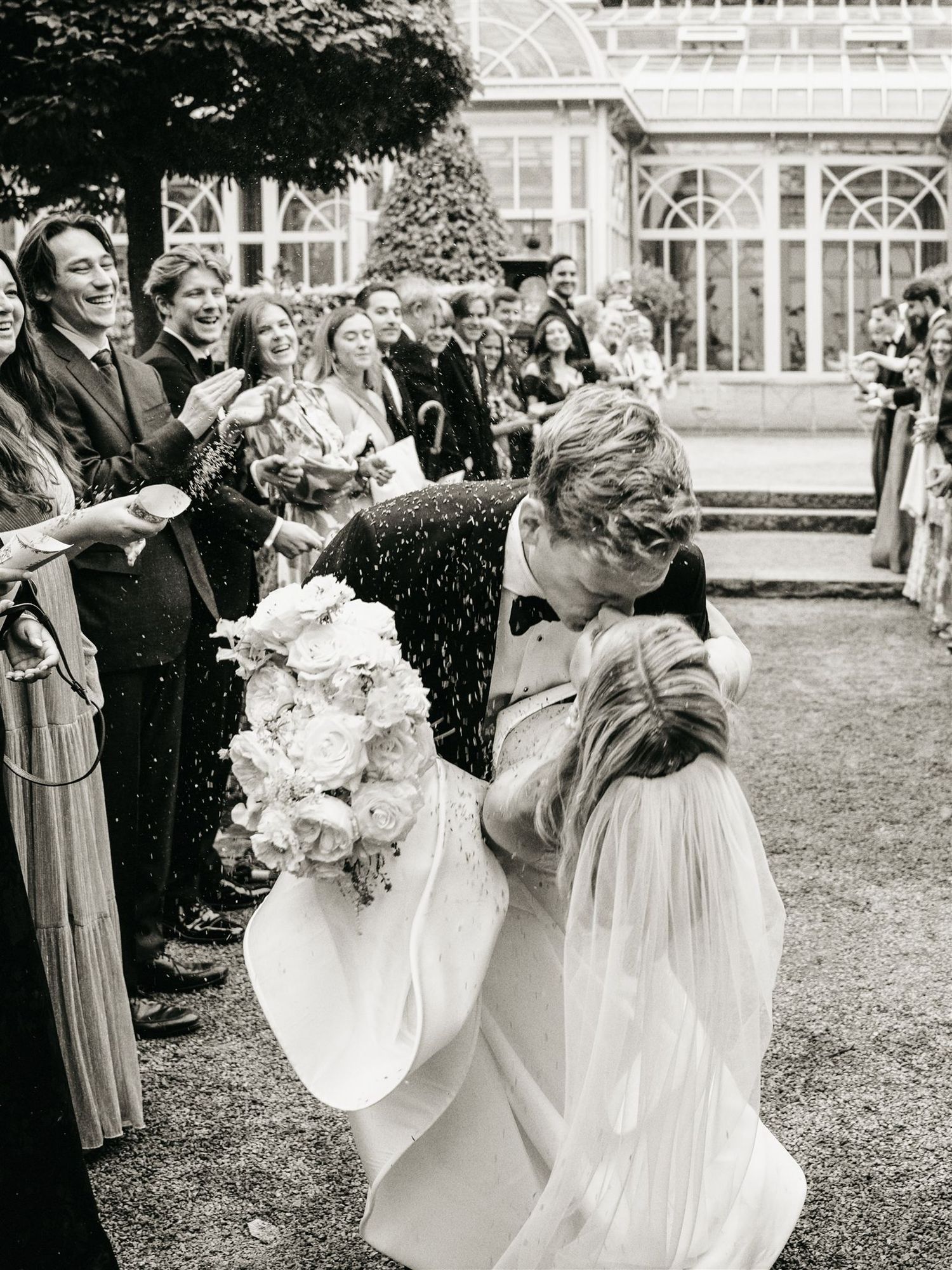 Groom kissing bride as guests celebrate with confetti outside a glass greenhouse in black and white.
