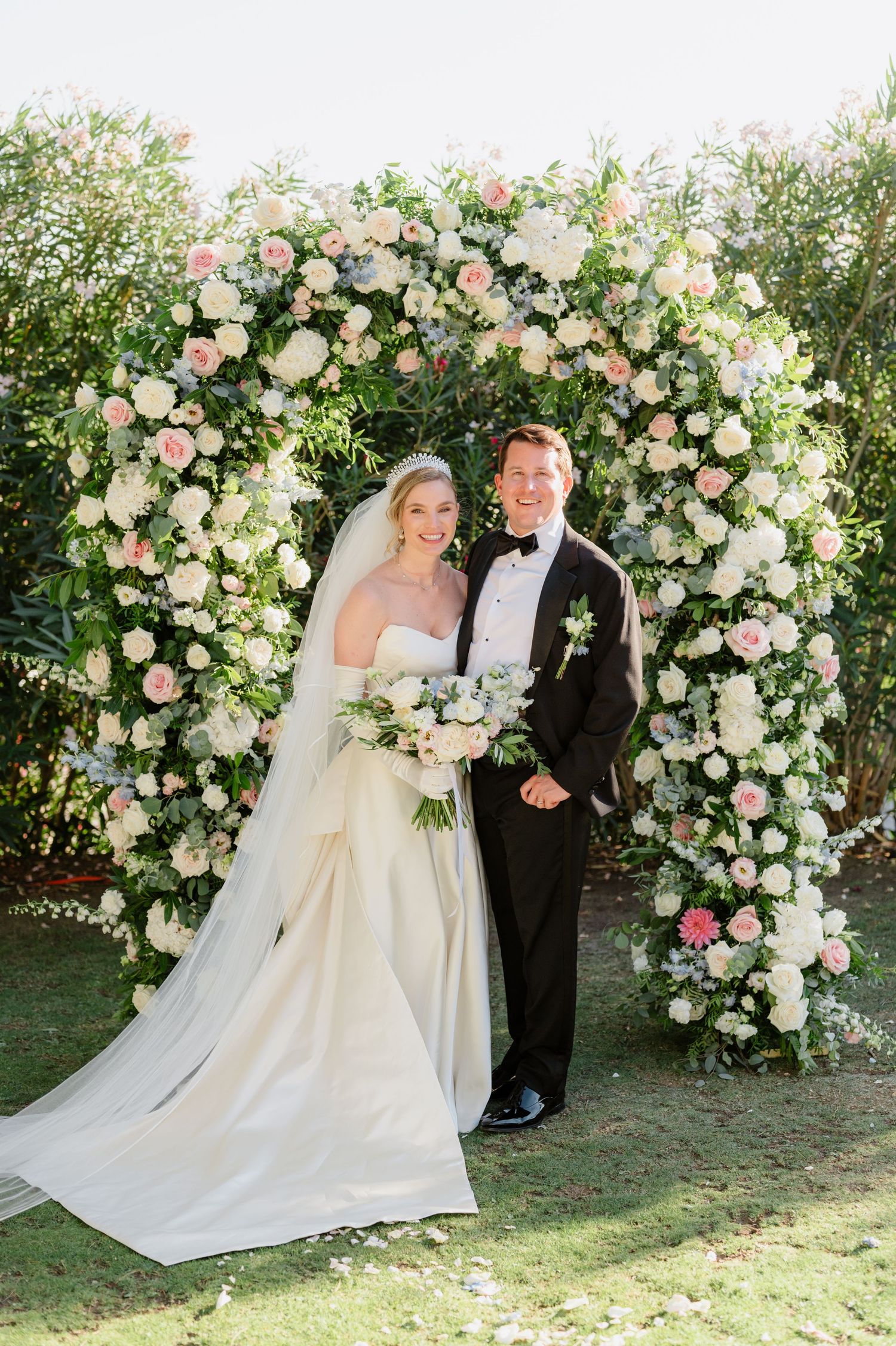 Elegant wedding couple poses in front of a lush floral arch with pink and white roses in a garden setting.