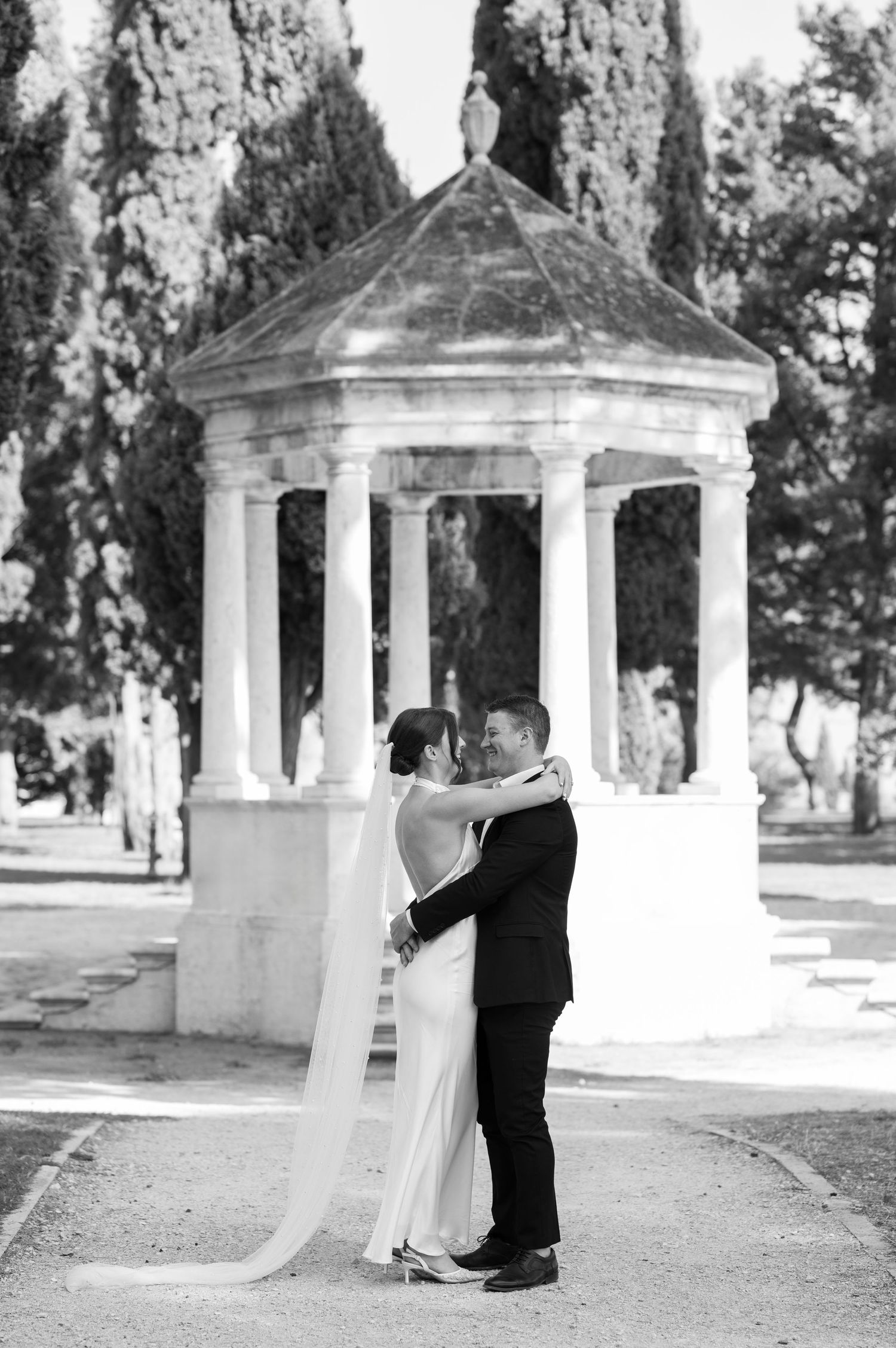 A romantic wedding couple embraces under an elegant white stone gazebo in a cypress-lined garden setting.