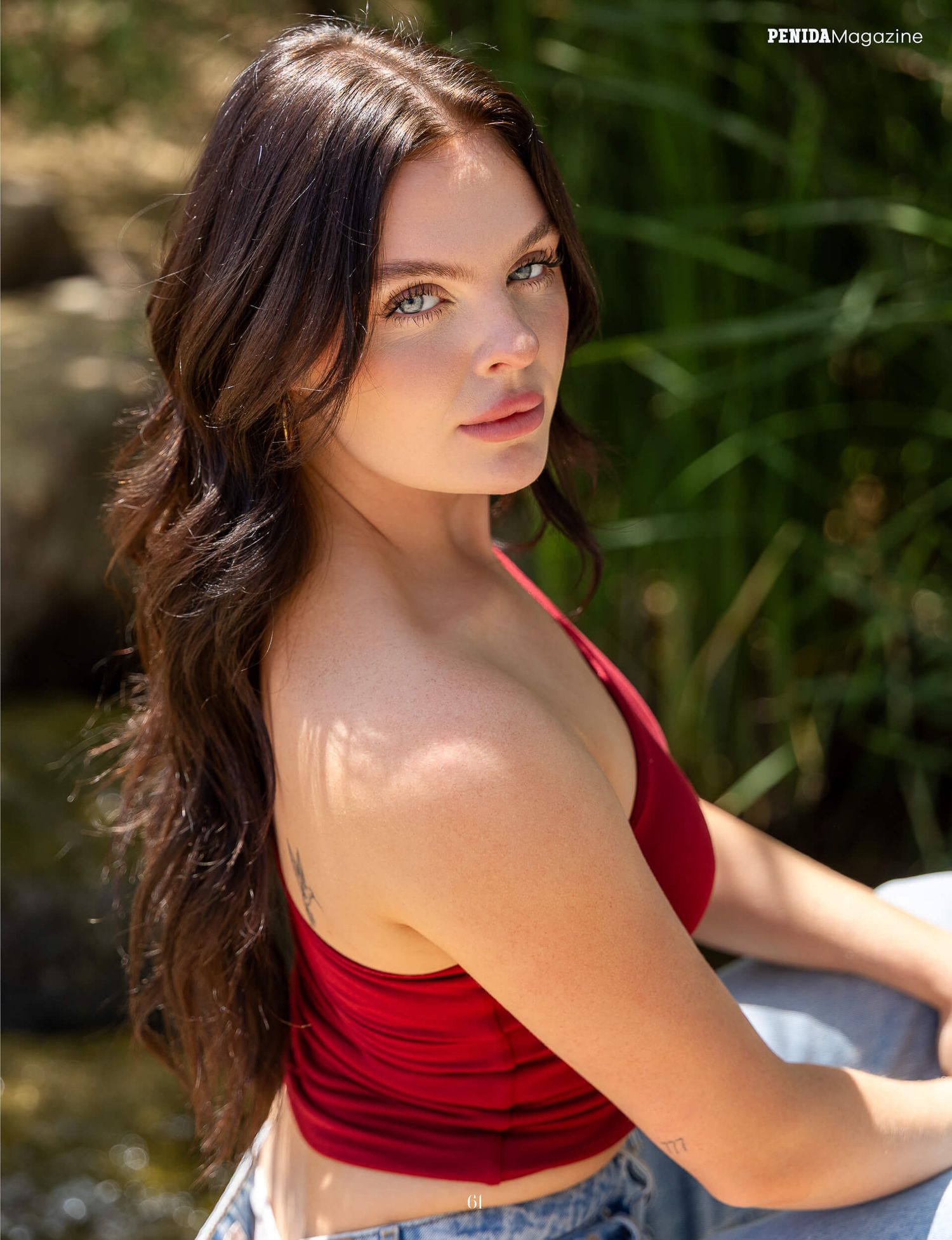 Brunette woman with green eyes in red crop top poses outdoors in natural sunlight.