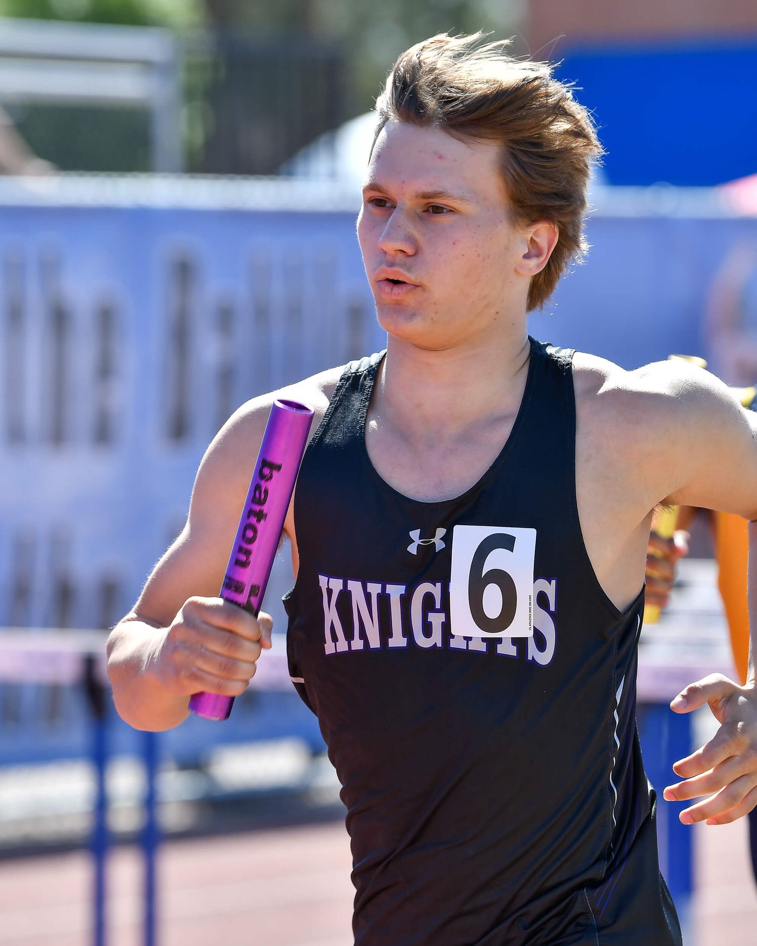 A young male relay runner in a Knights jersey holds a baton during a track and field race.