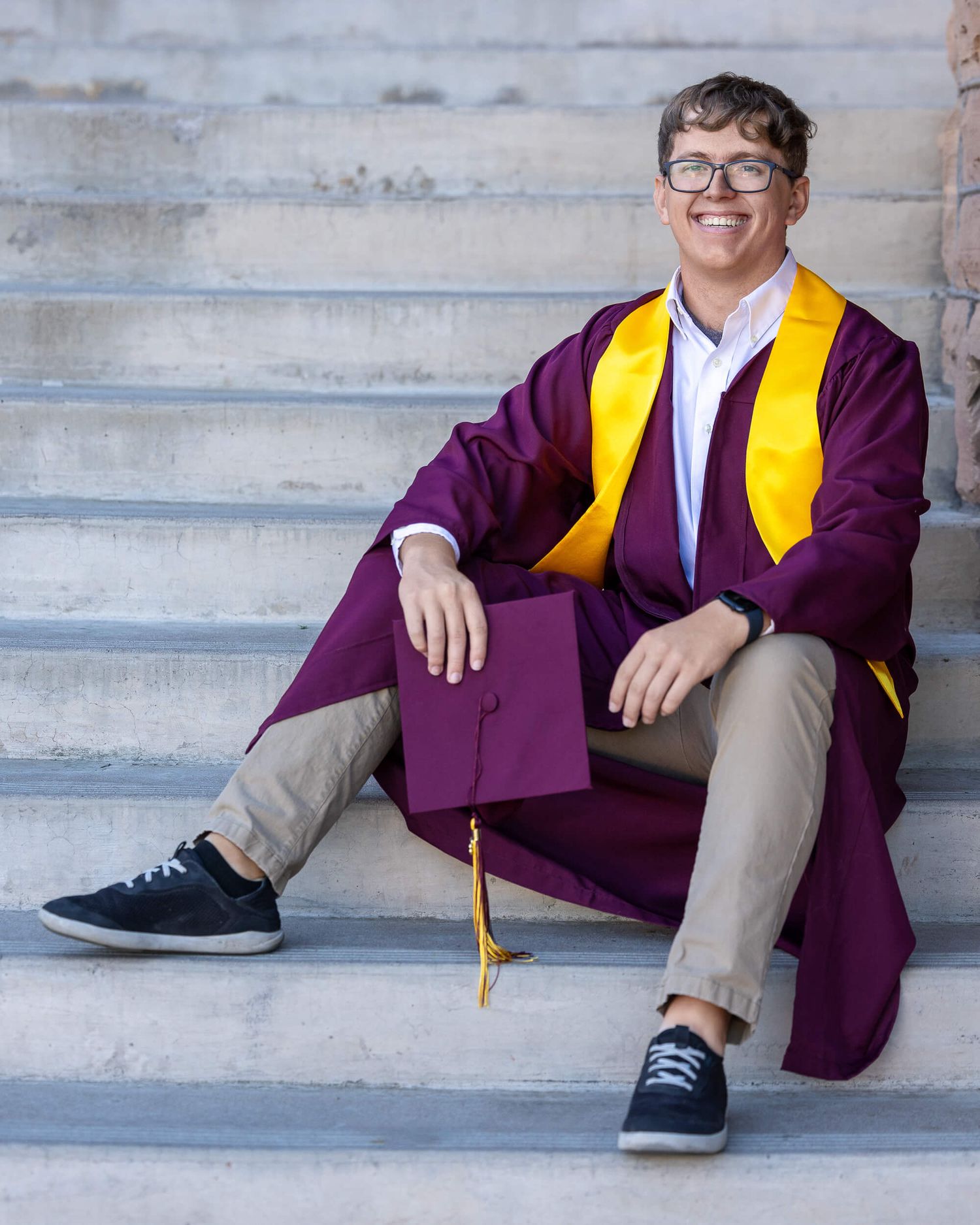 Young man in maroon and gold graduation regalia sitting on steps holding cap.