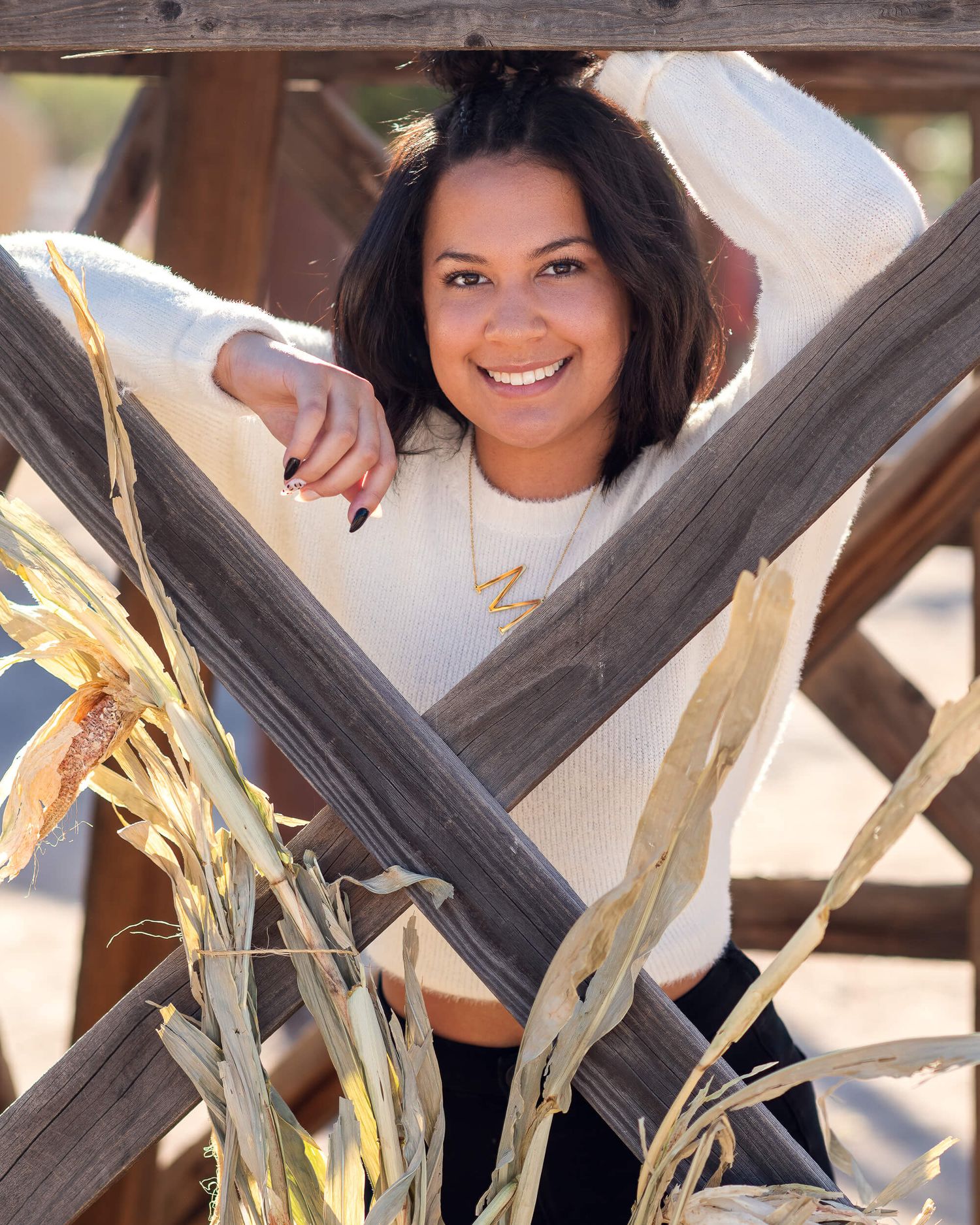 Smiling young woman in a white sweater poses between rustic wooden beams with dried corn stalks in an outdoor setting.