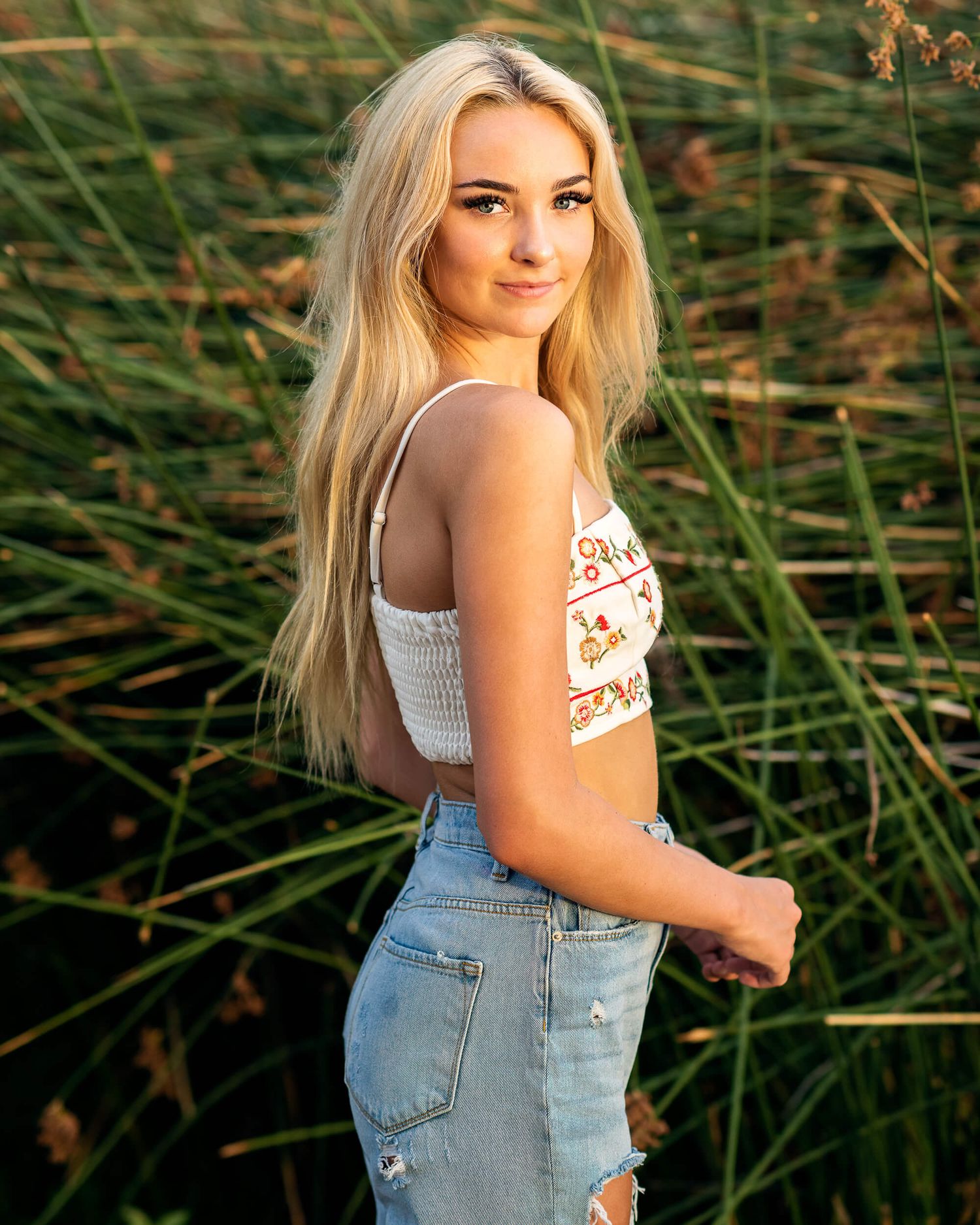 Young blonde woman in crop top and jeans posing outdoors in tall green grass during golden hour.