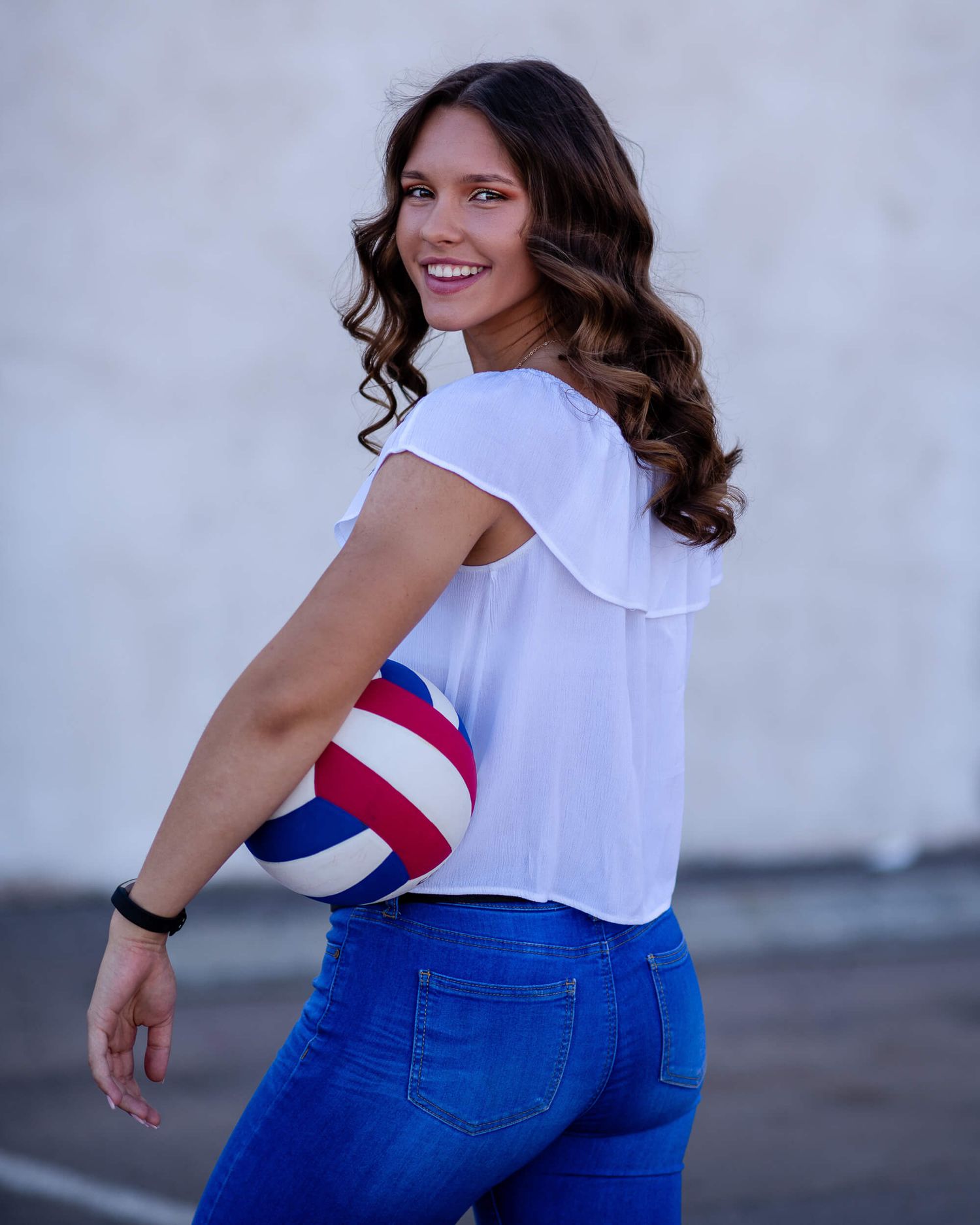 Smiling brunette woman in white crop top and blue jeans holding a red, white, and blue volleyball outdoors.