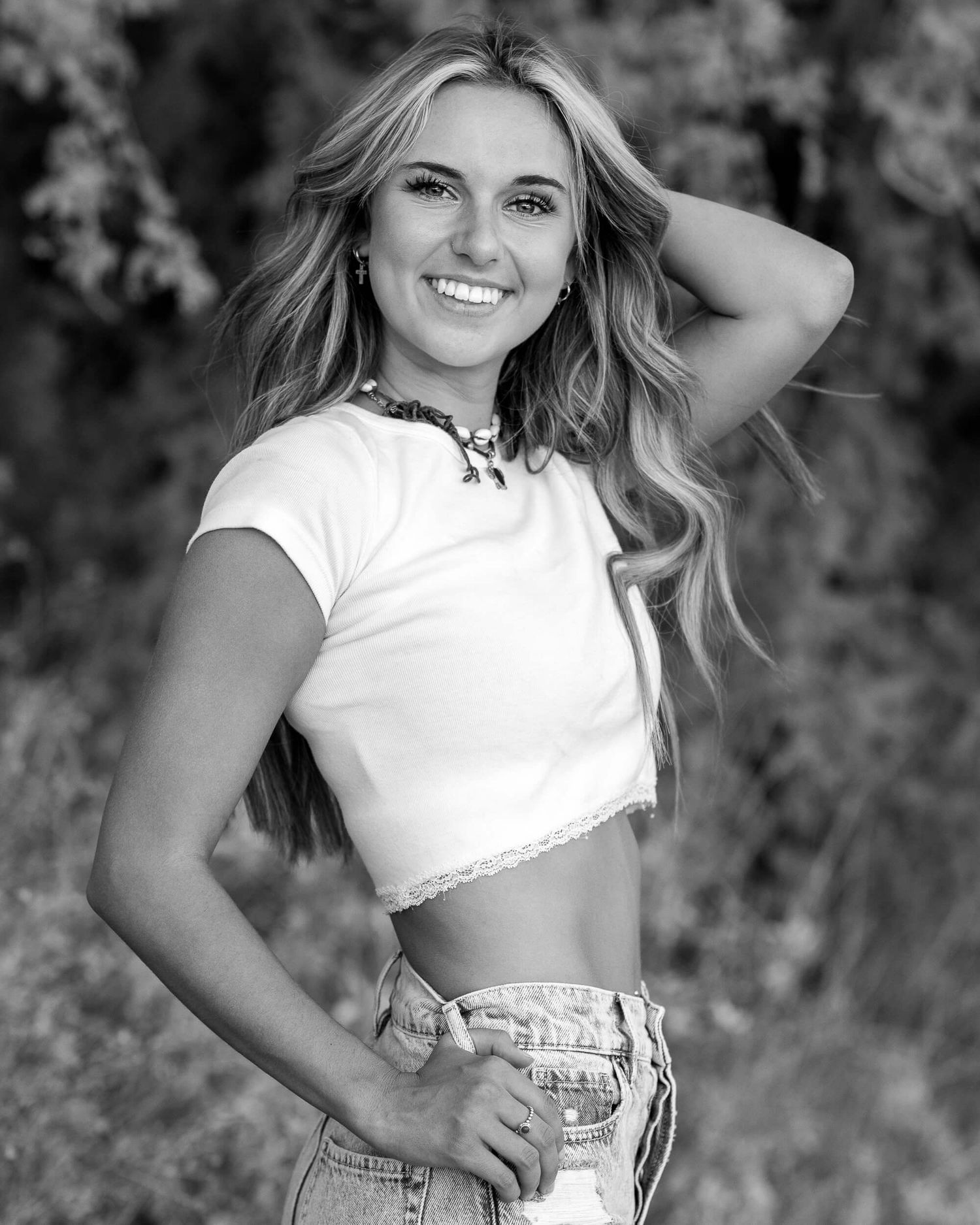 Black and white photo of a smiling blonde woman in a white crop top and jeans posing outdoors.