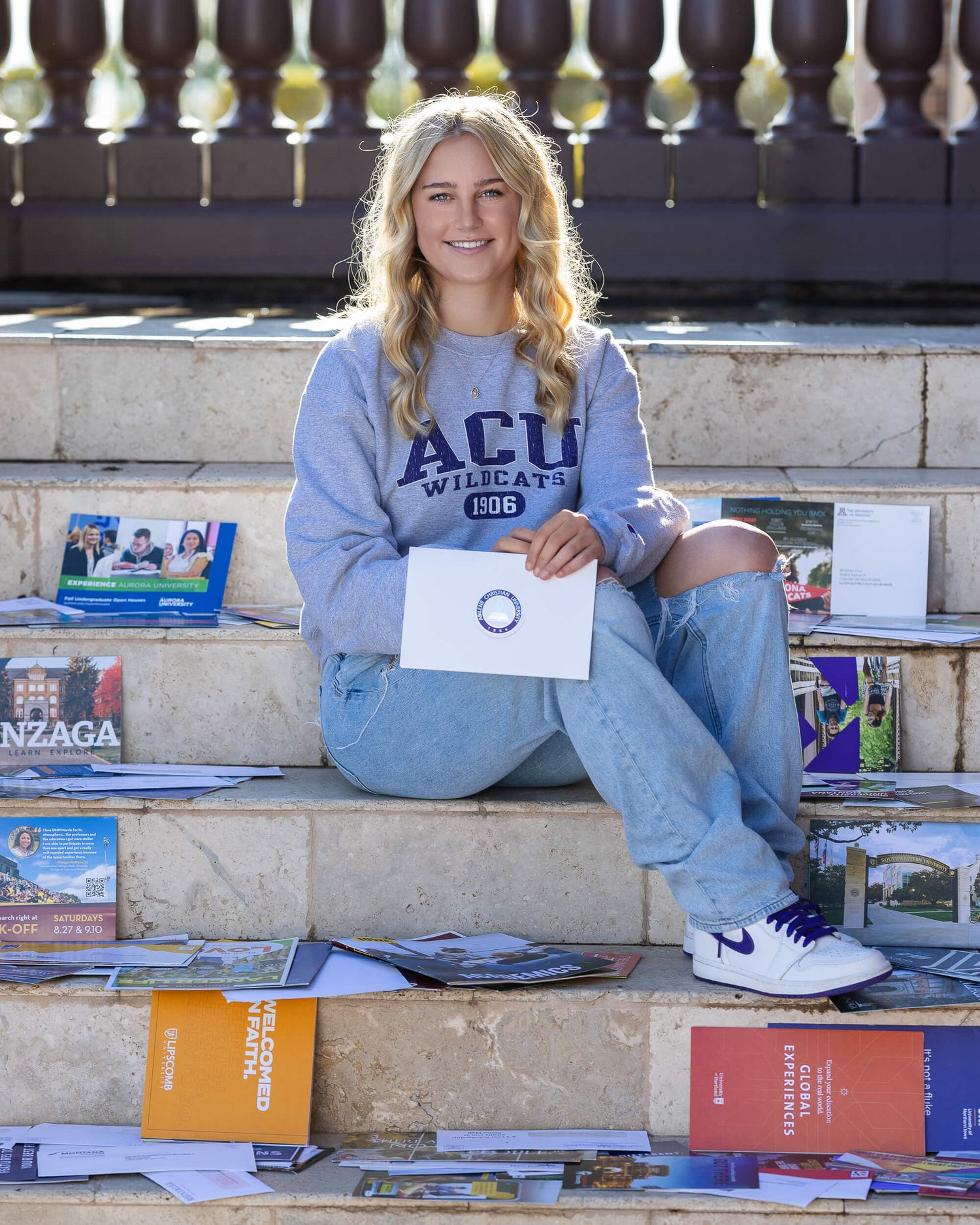 Blonde teen girl sitting on steps surrounded by college brochures wearing ACU sweatshirt.