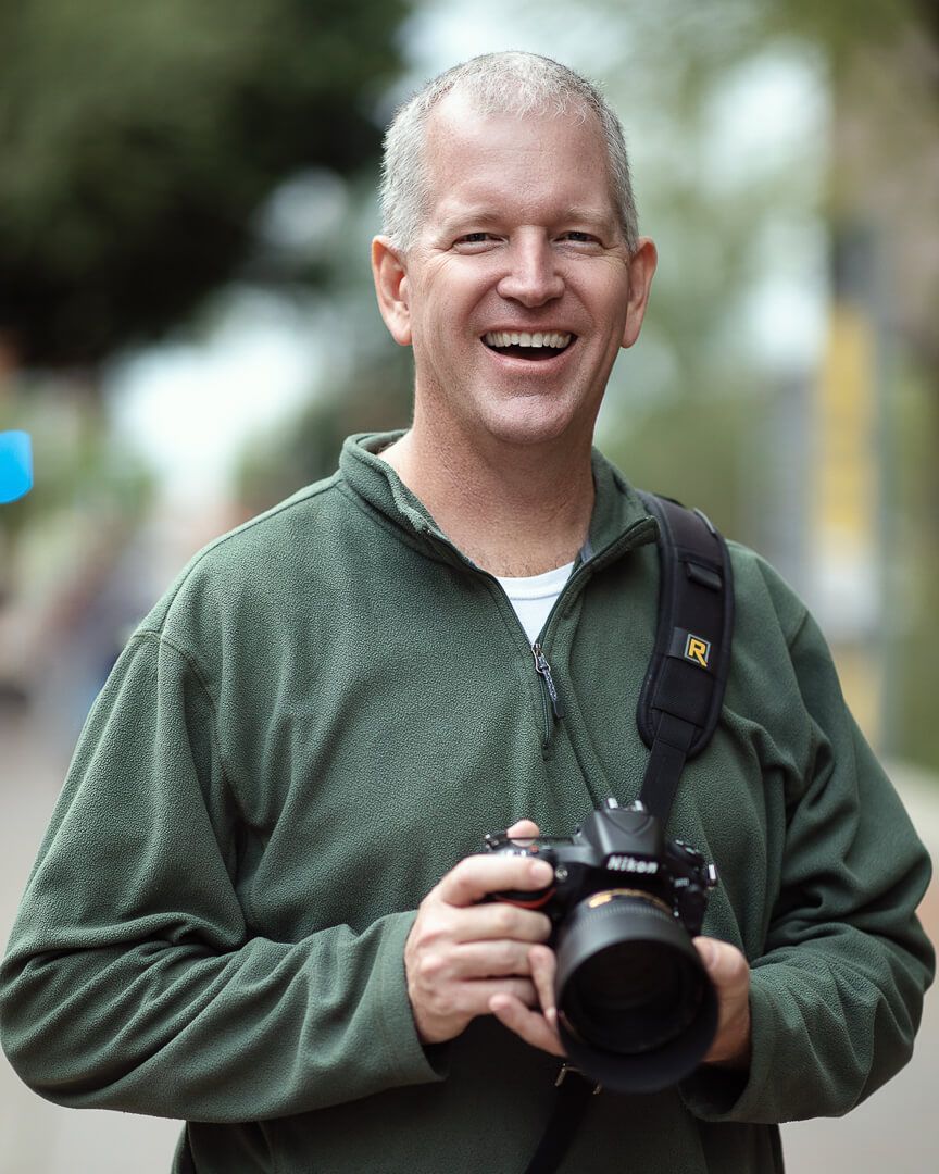 Smiling man in green fleece jacket holding a Nikon camera outdoors with a blurred street background.