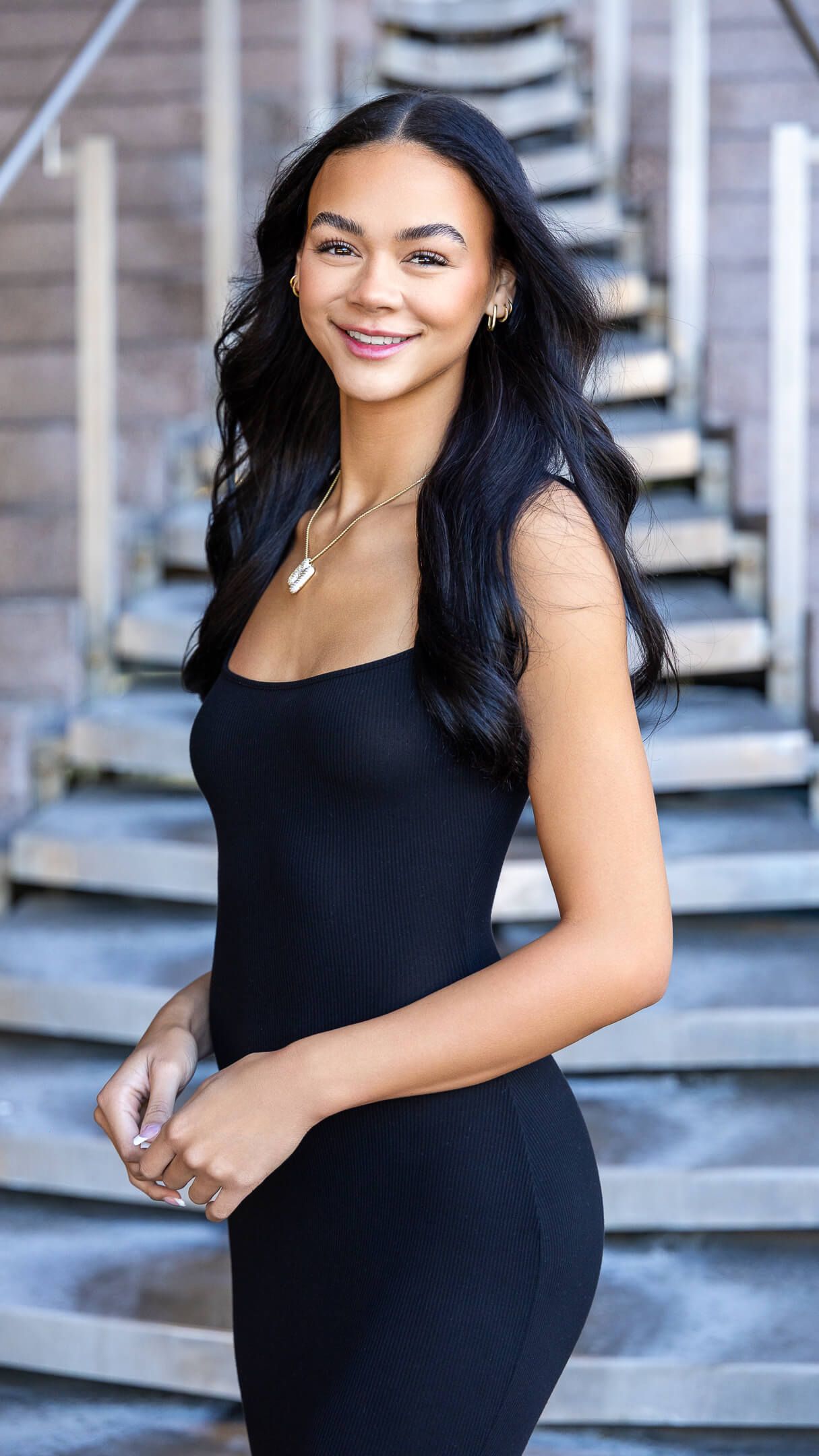 Young woman in black fitted dress smiling outdoors near stone staircase with long dark wavy hair.
