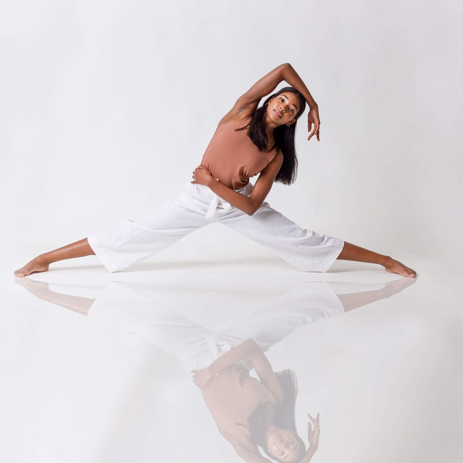 Flexible woman in rust top and white pants doing a split stretch in a white studio.