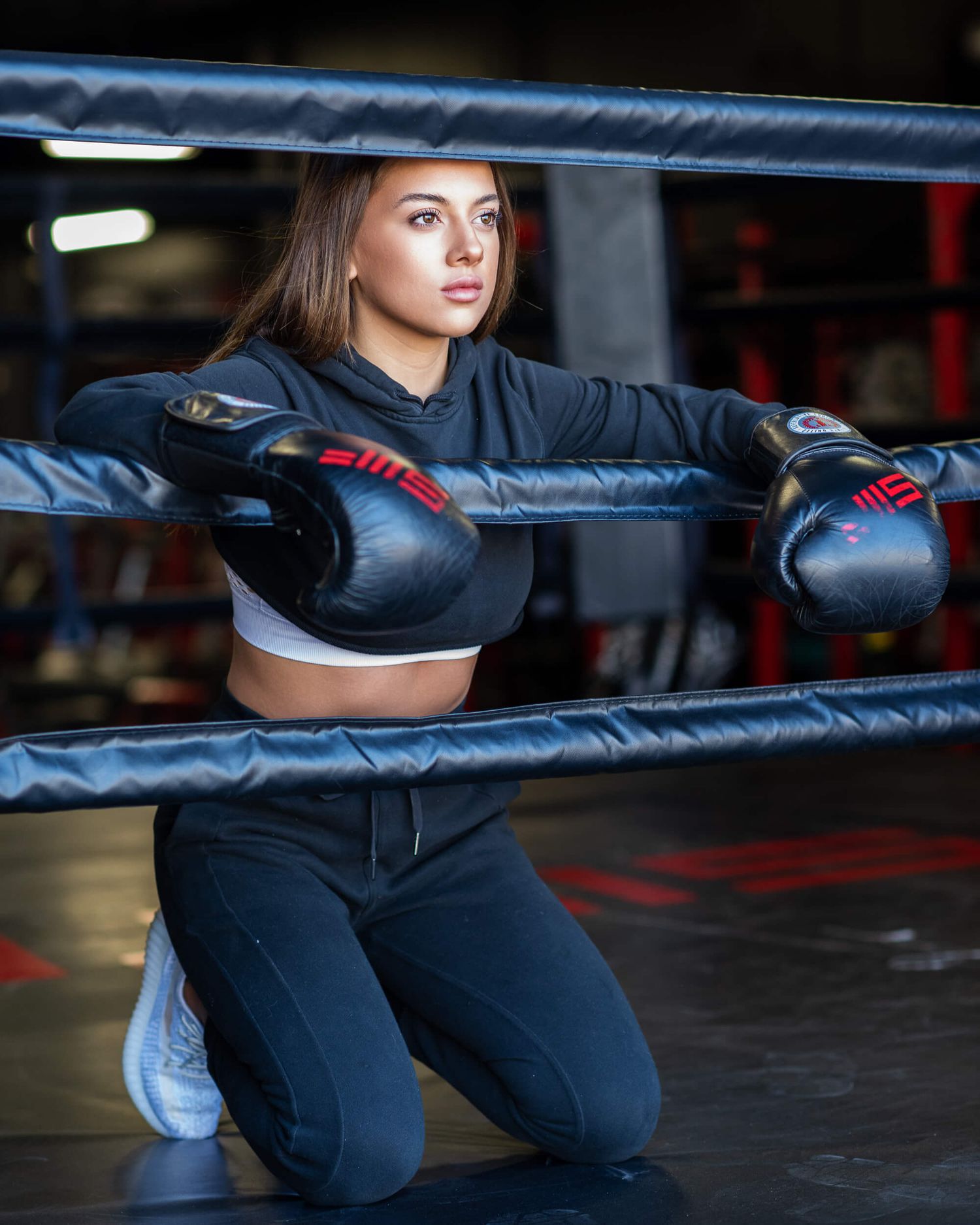 Female boxer in black gear kneeling against boxing ring ropes in a gym setting.