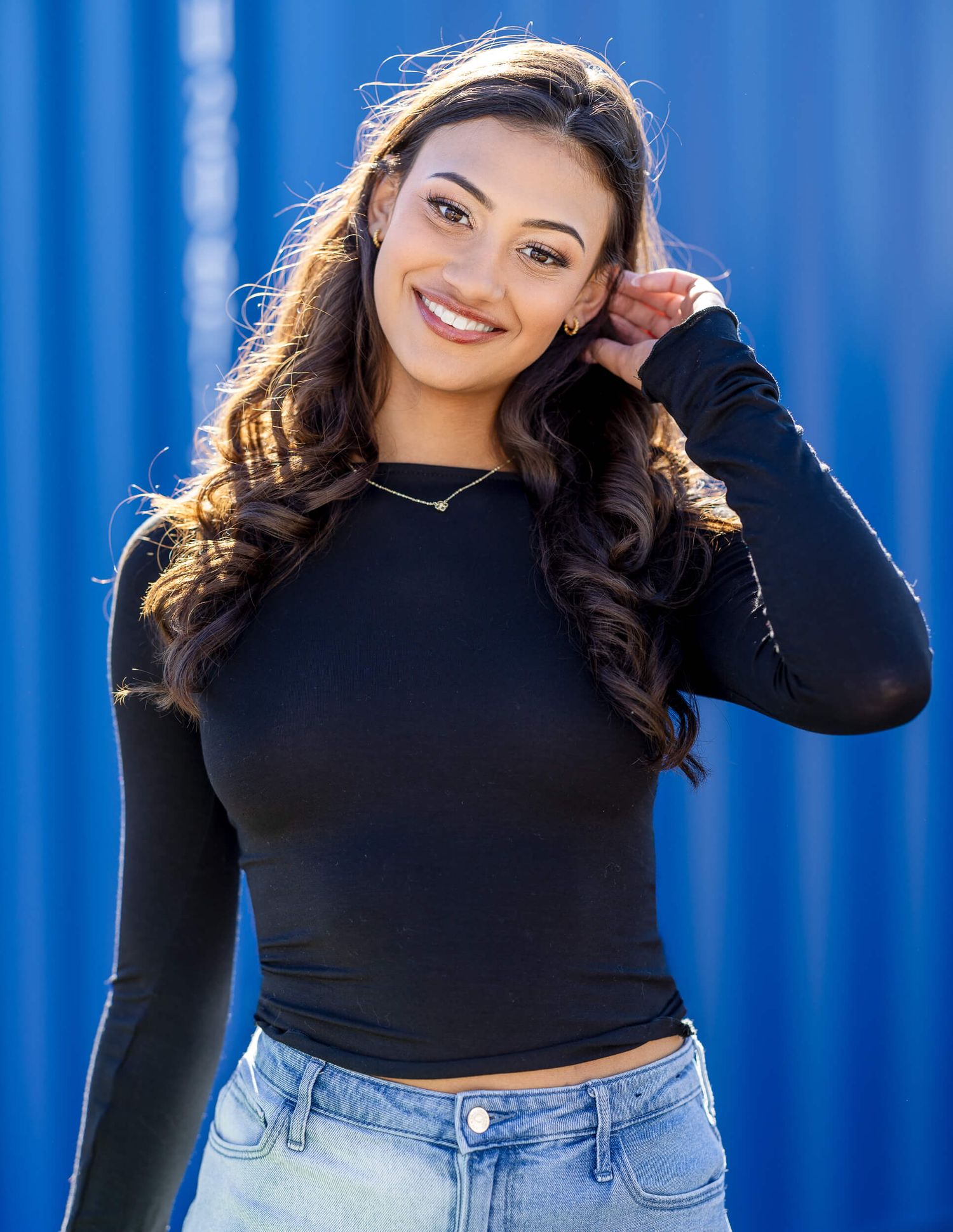 Smiling brunette woman in black long sleeve crop top against blue wall background.