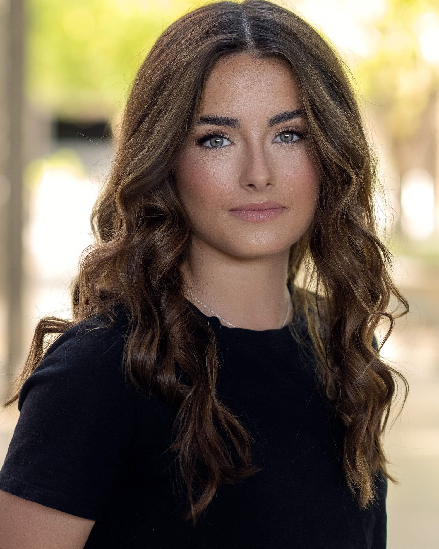 Young woman with long wavy brown hair and blue eyes in a black top with blurred outdoor background.