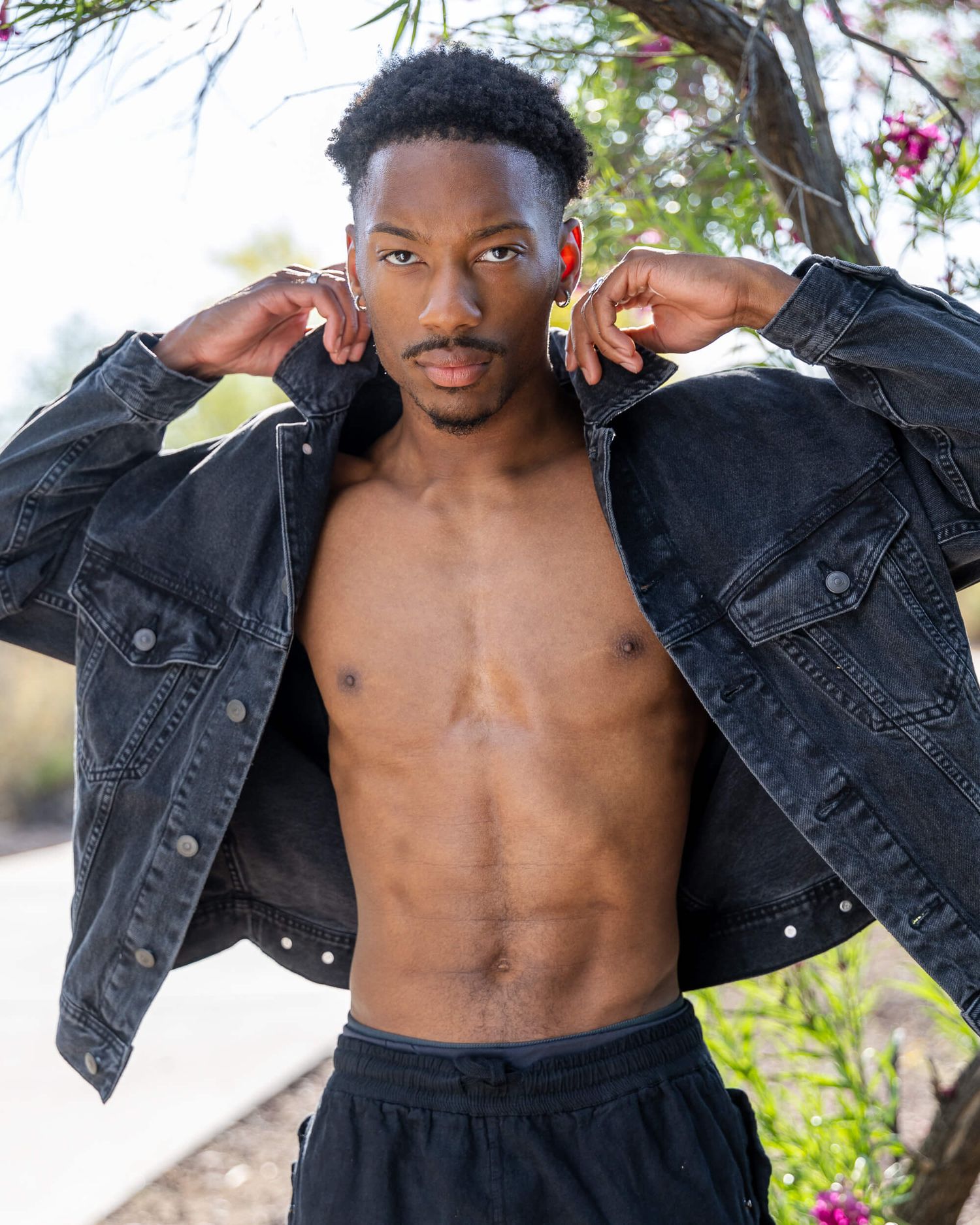 Shirtless young Black man opening black denim jacket outdoors near flowering trees and shrubs.