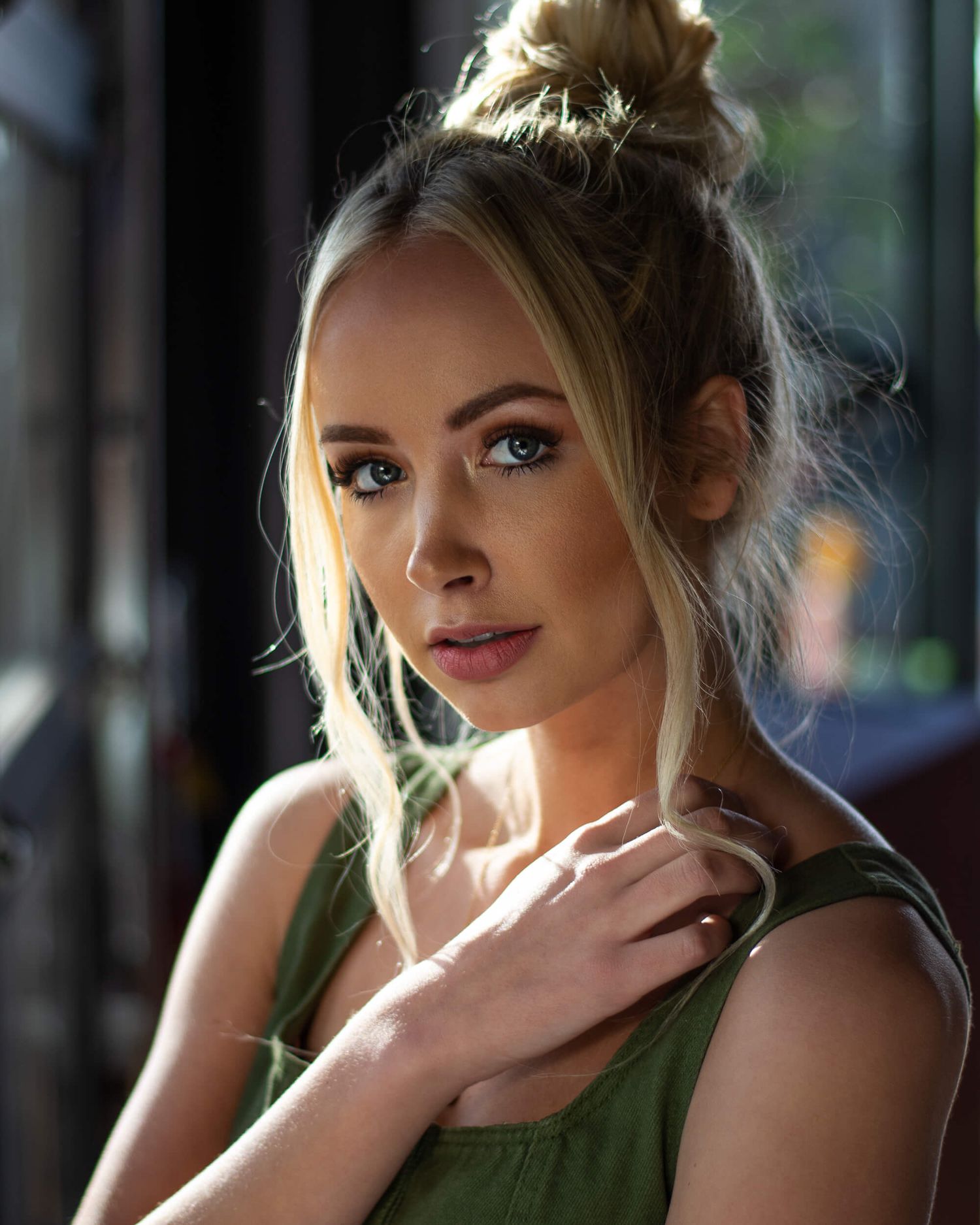 Blonde woman with green eyes in olive top with hair in bun posing indoors.