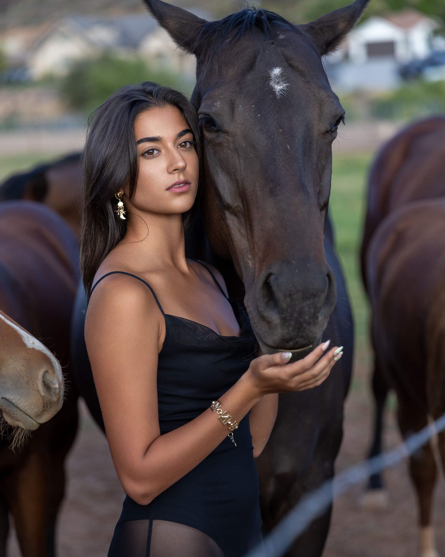 Dark-haired woman in black bodysuit feeding a dark horse at a ranch with horses behind.