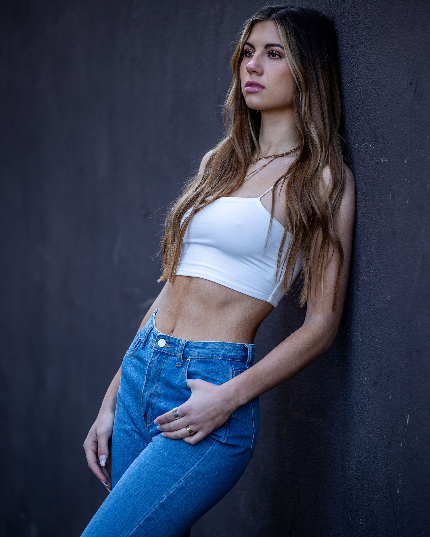 Young woman in white crop top and blue jeans leaning against dark wall.