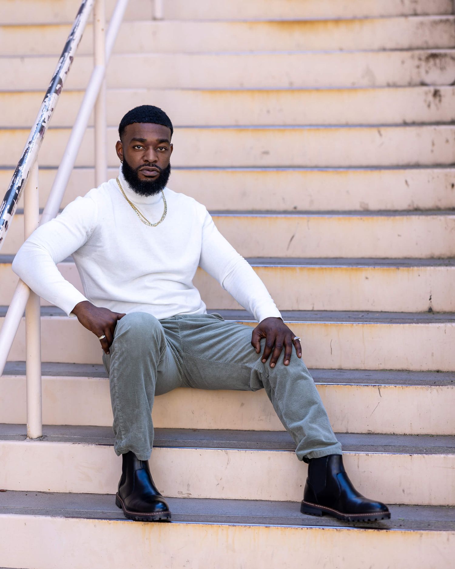 Young Black man in white sweater, olive pants, and black boots sitting on outdoor stairs.