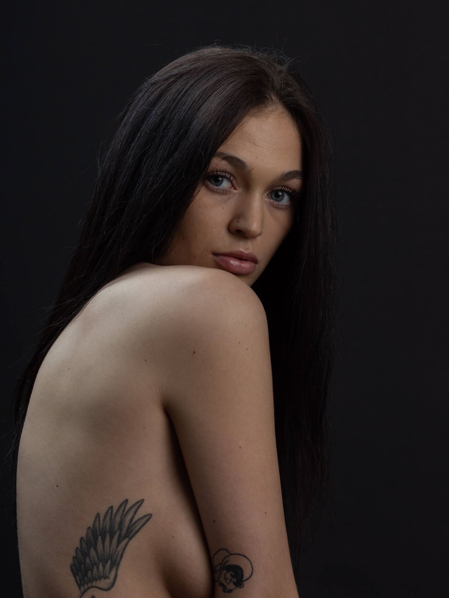 Young woman with long dark hair and arm tattoos poses against dark background in artistic portrait.