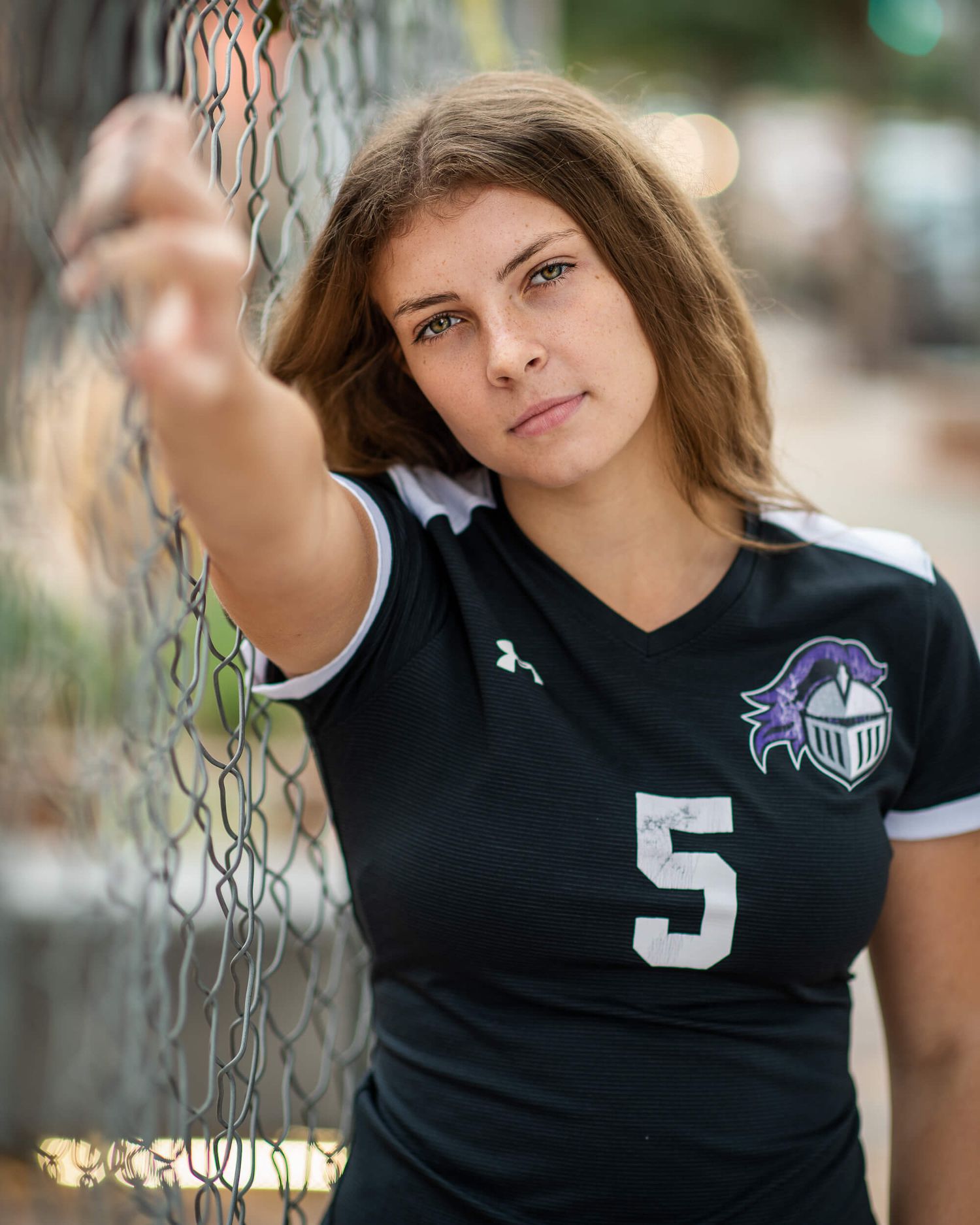 Athletic young woman in soccer jersey number 5 reaching toward camera near chain-link fence.