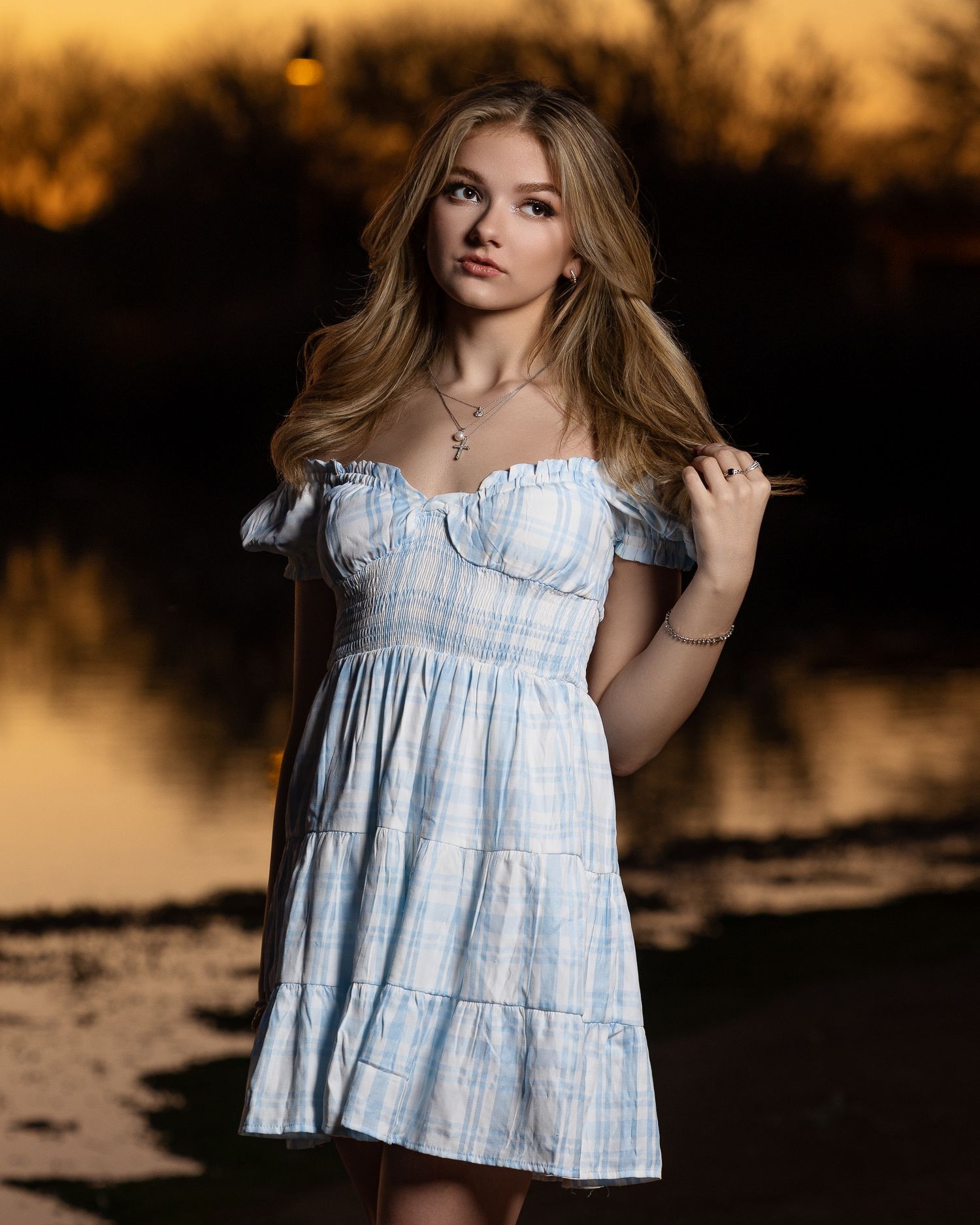 Young blonde woman in blue off-shoulder dress poses near lake at golden sunset.