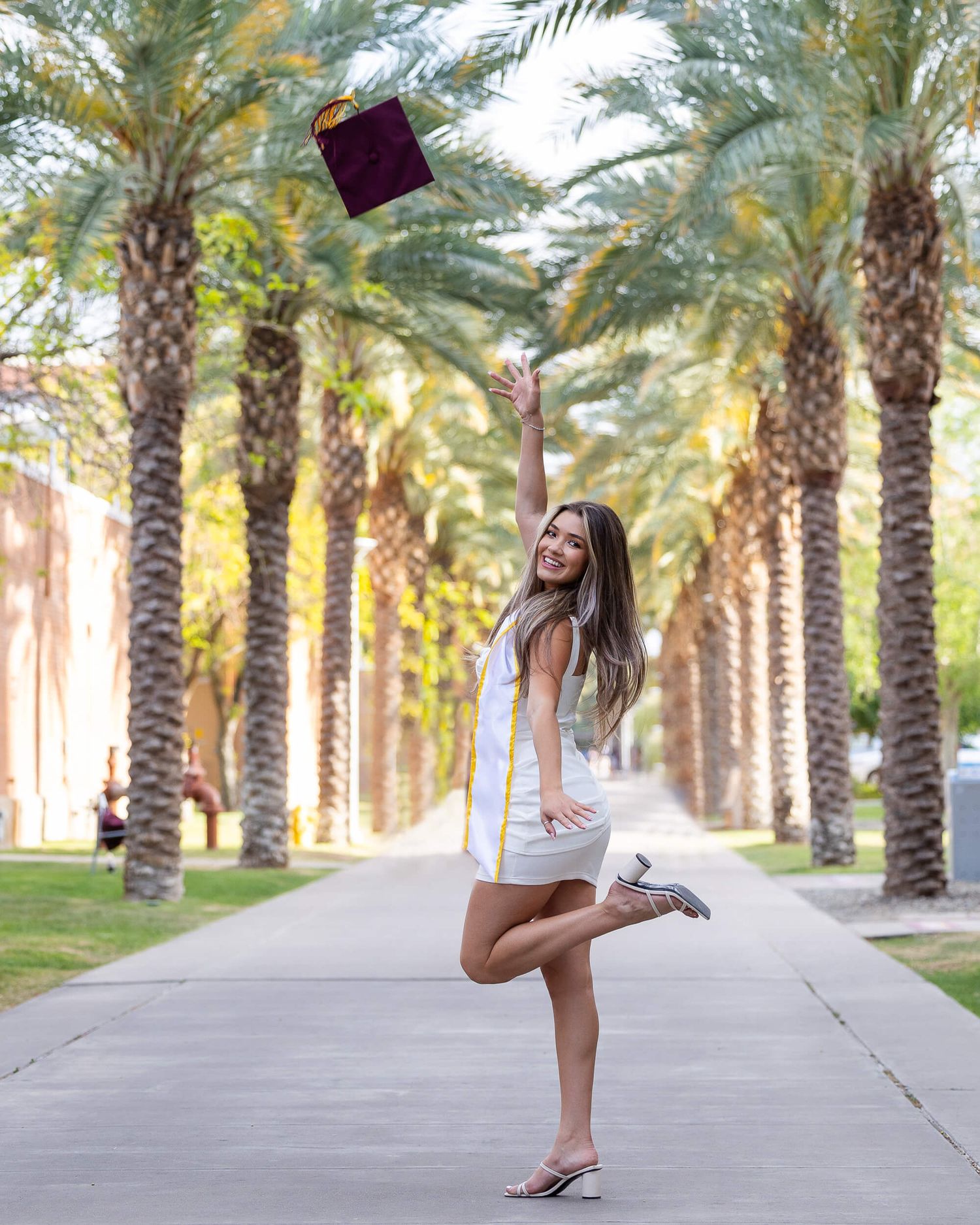A graduate tosses her maroon cap in the air along a sunny palm tree-lined campus path.