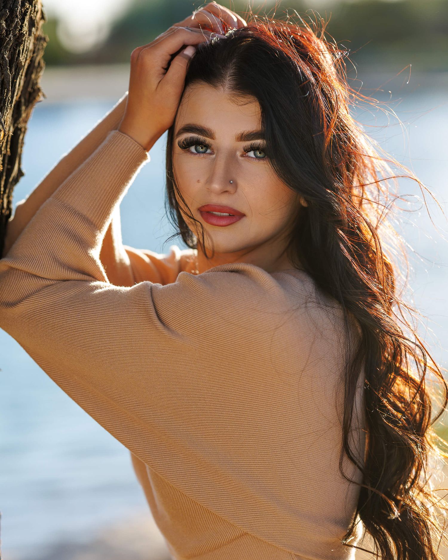 Dark-haired woman in beige sweater poses by waterside in warm golden light.
