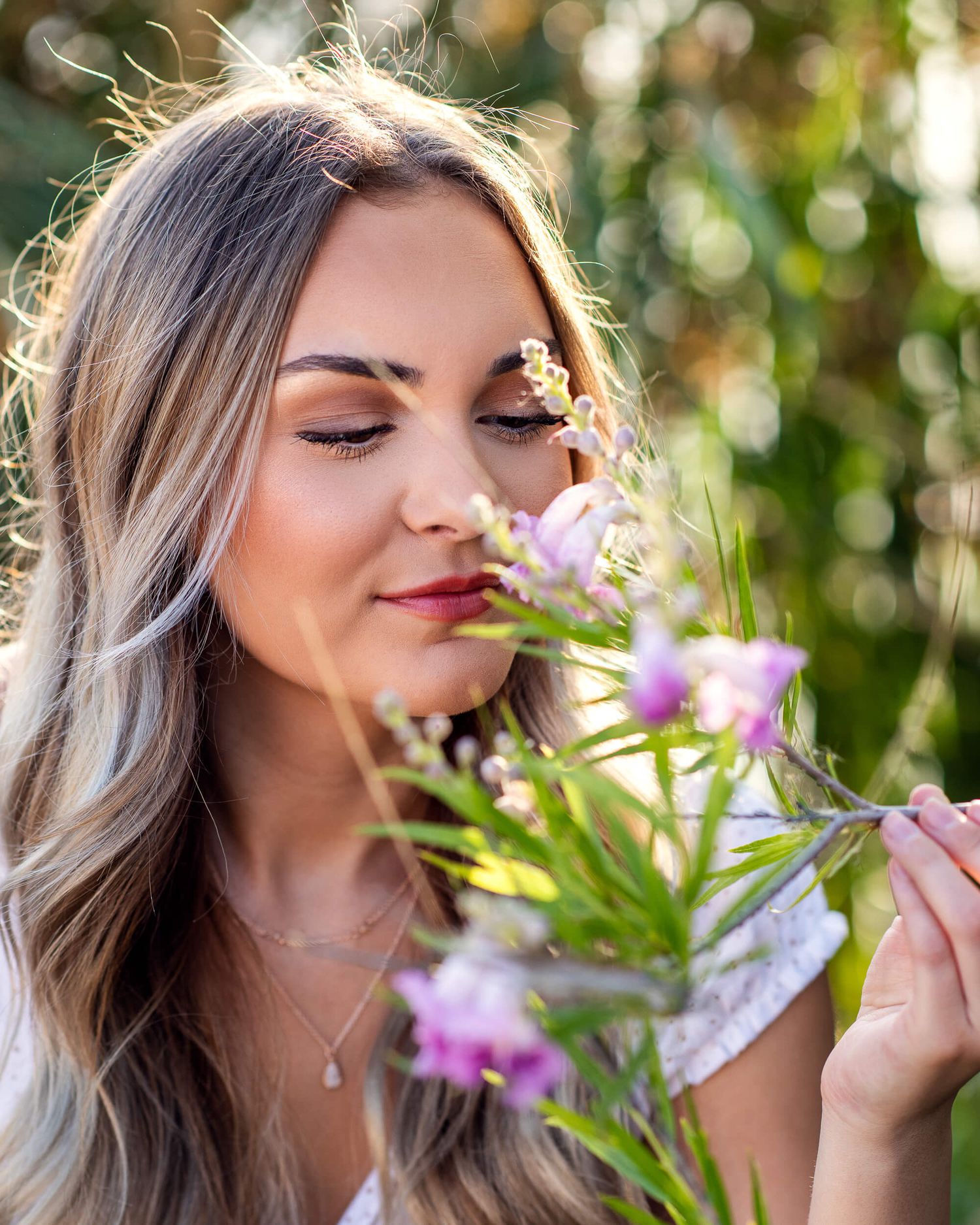 Young blonde woman with eyes closed smells pink wildflowers in a sunny garden.