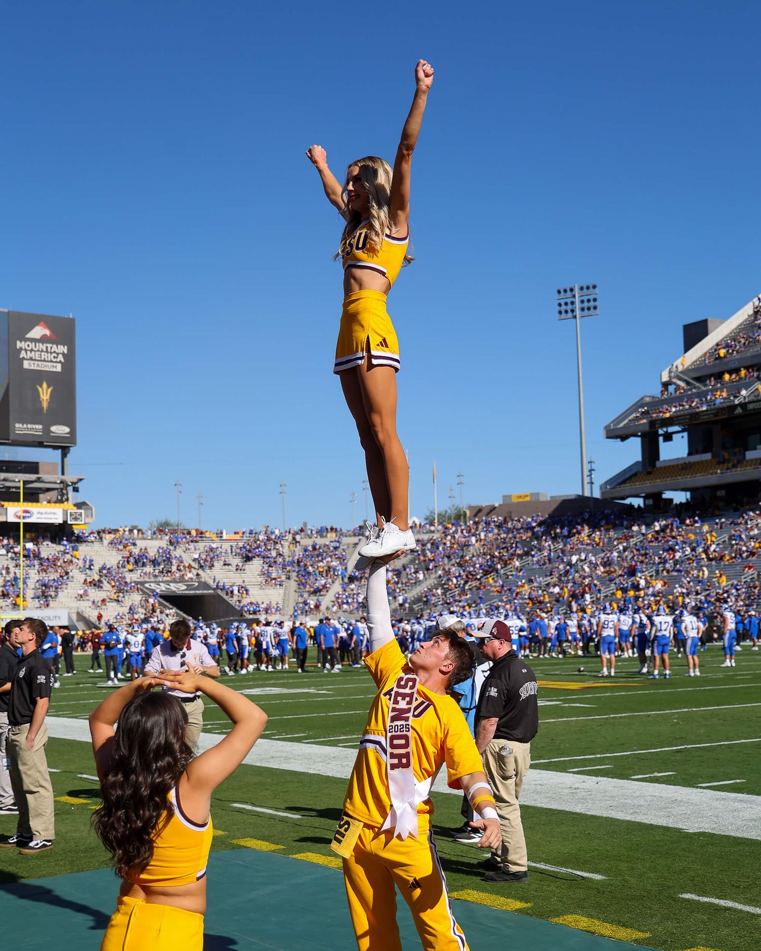ASU cheerleader in gold uniform performs a liberty stunt at a sunny college football game.