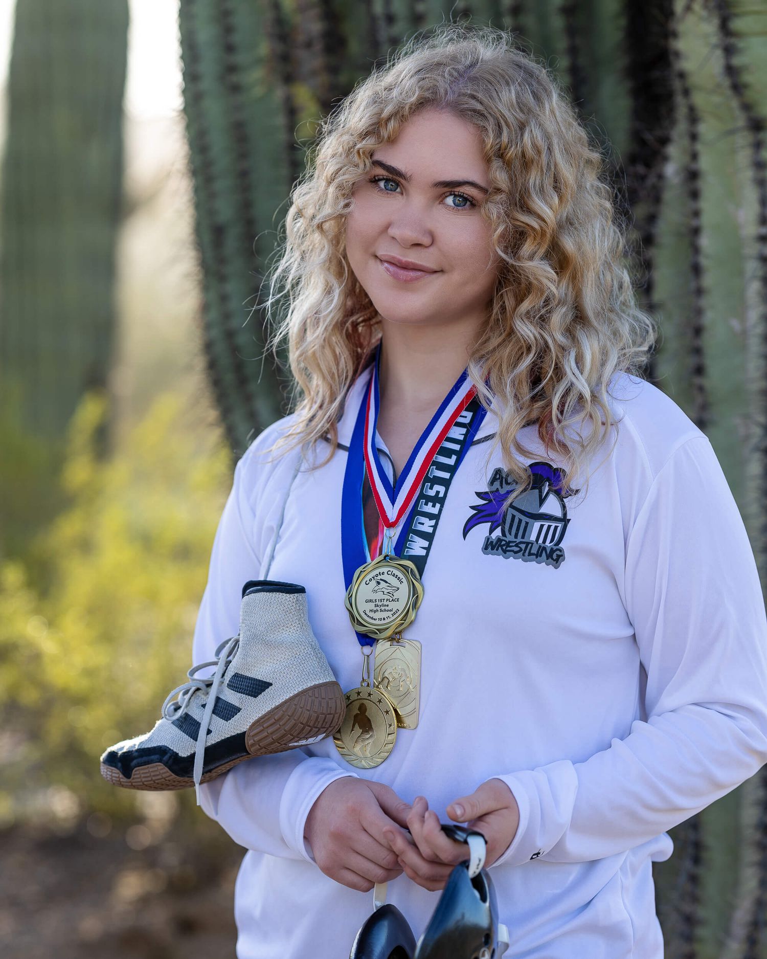 A young female wrestler poses with gold medals and wrestling shoes near a saguaro cactus.