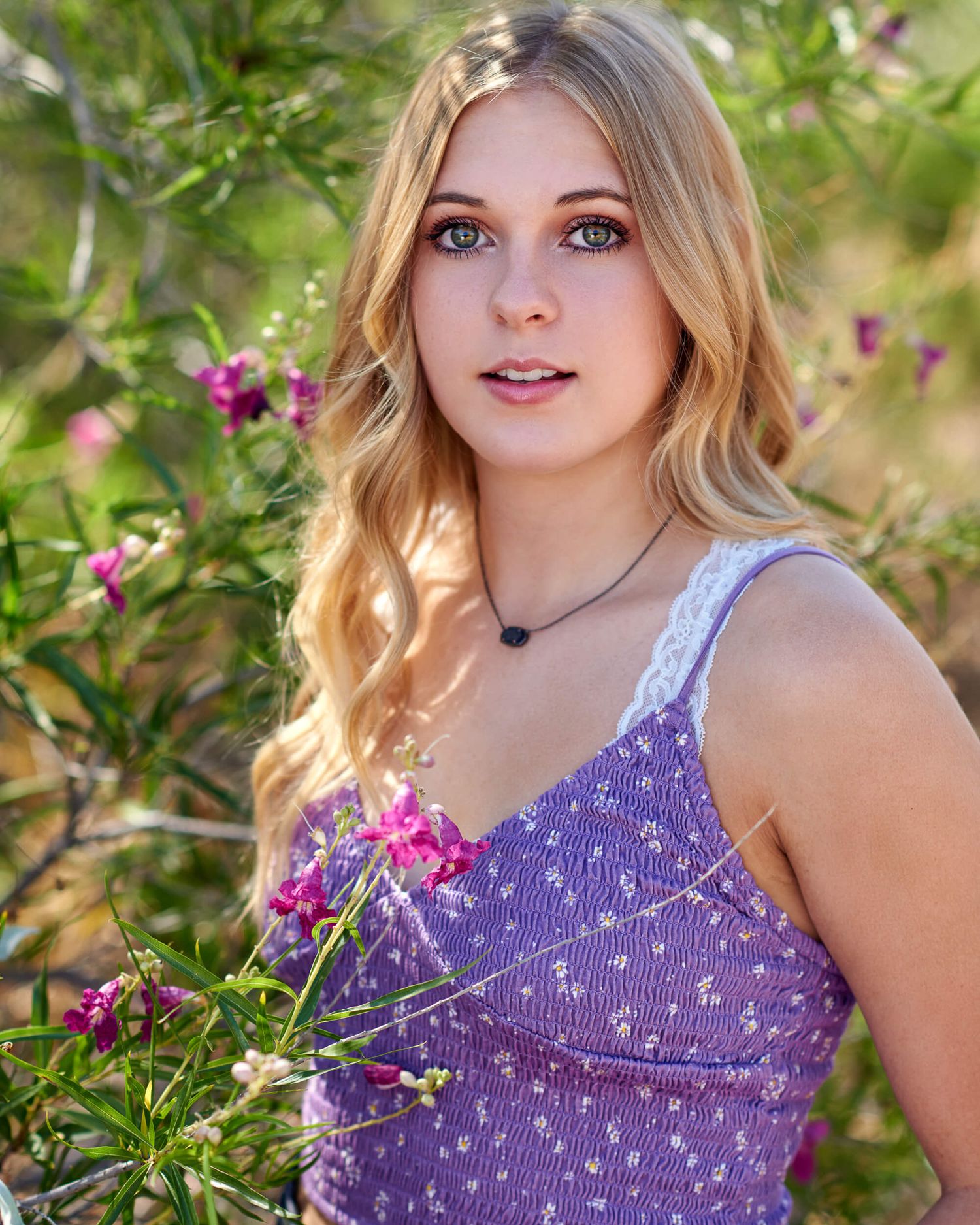 Blonde teenage girl with blue eyes in purple floral top posing near pink blooming flowers.