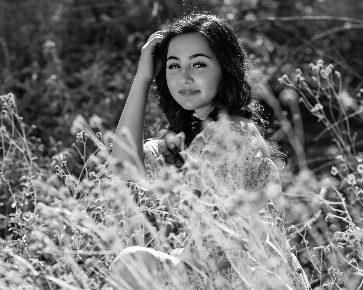 Black and white portrait of a young woman sitting among wildflowers, hand in hair, smiling softly.