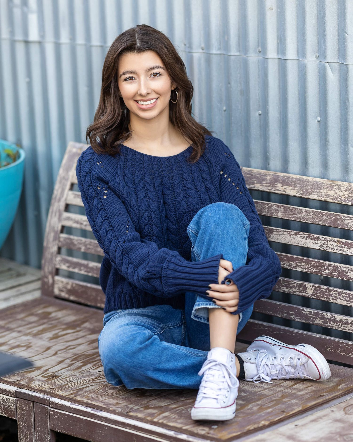 Young woman in navy cable-knit sweater and jeans sitting cross-legged on wooden bench, smiling outdoors.