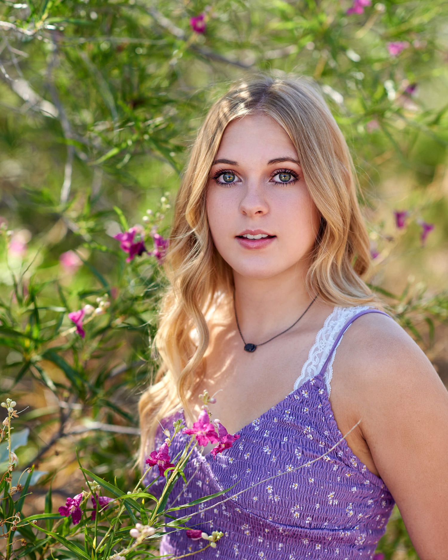 Young blonde woman in purple floral dress poses among pink blooming flowers in a lush green garden.